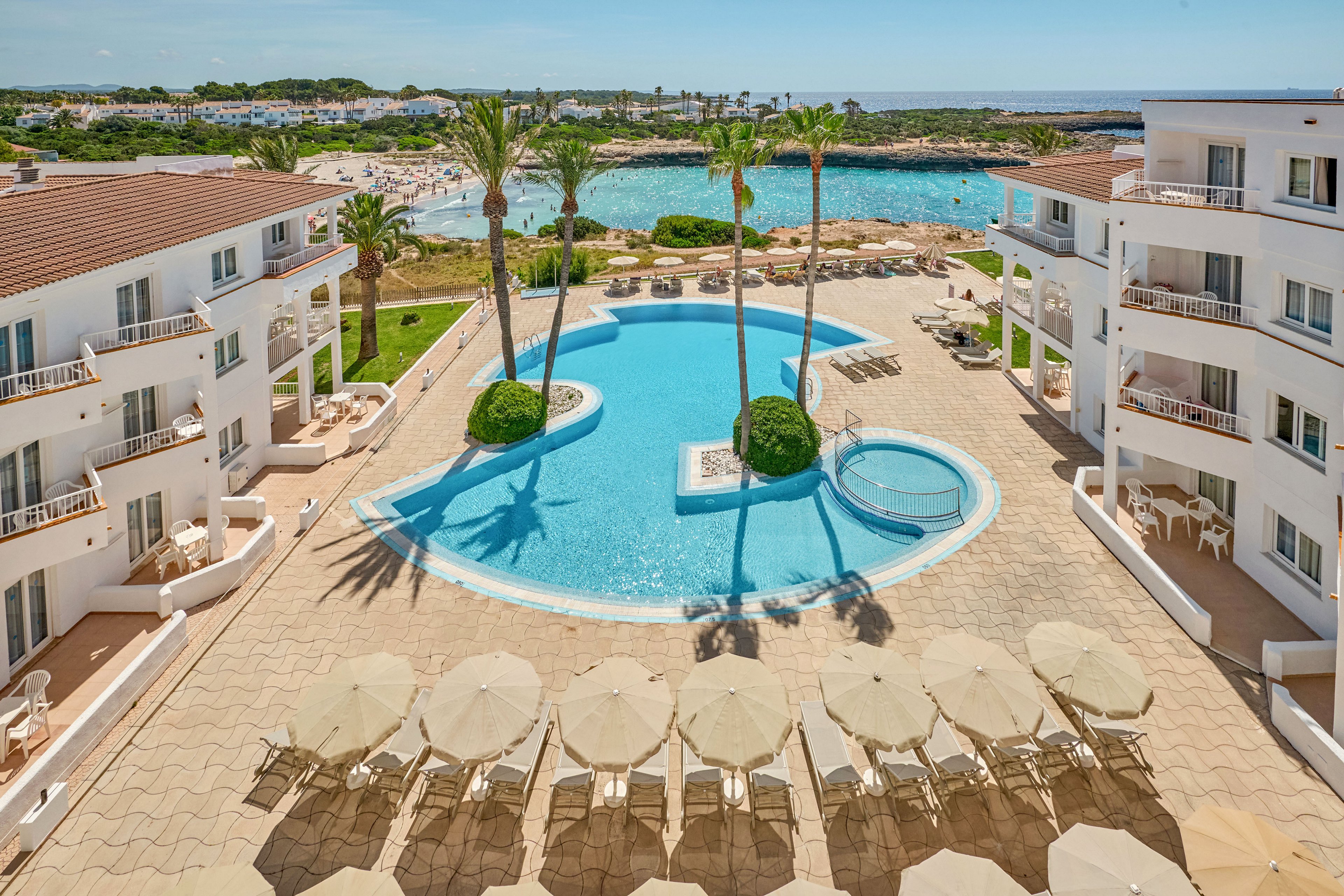A bright seaside resort features a central outdoor swimming pool surrounded by white apartment-style buildings. Sun loungers and umbrellas line the tiled terrace, creating a relaxed holiday atmosphere. Tall palm trees rise from landscaped islands within the pool, and a small circular pool adjoins the main one. In the background, a calm blue bay and rocky shoreline complete the scenic coastal setting under a clear sky.