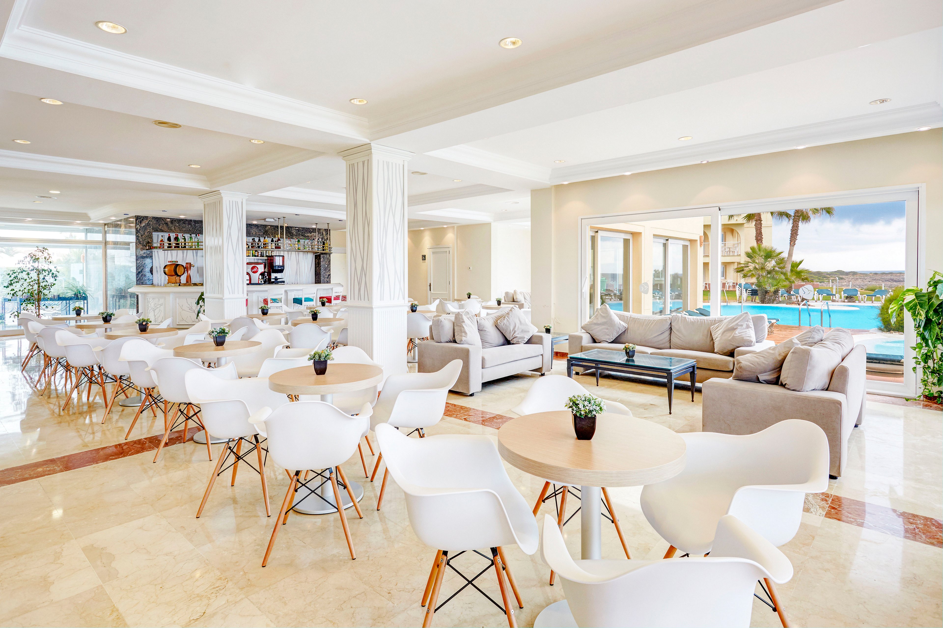 A spacious, contemporary lounge area features white chairs and round tables arranged neatly across a polished floor. Neutral-toned sofas with cushions create a comfortable seating zone near large windows and sliding doors. Beyond the glass, a turquoise swimming pool and palm trees overlook a scenic waterfront landscape. The space is brightly lit with natural light, giving it an airy, inviting atmosphere suitable for relaxation or casual dining.