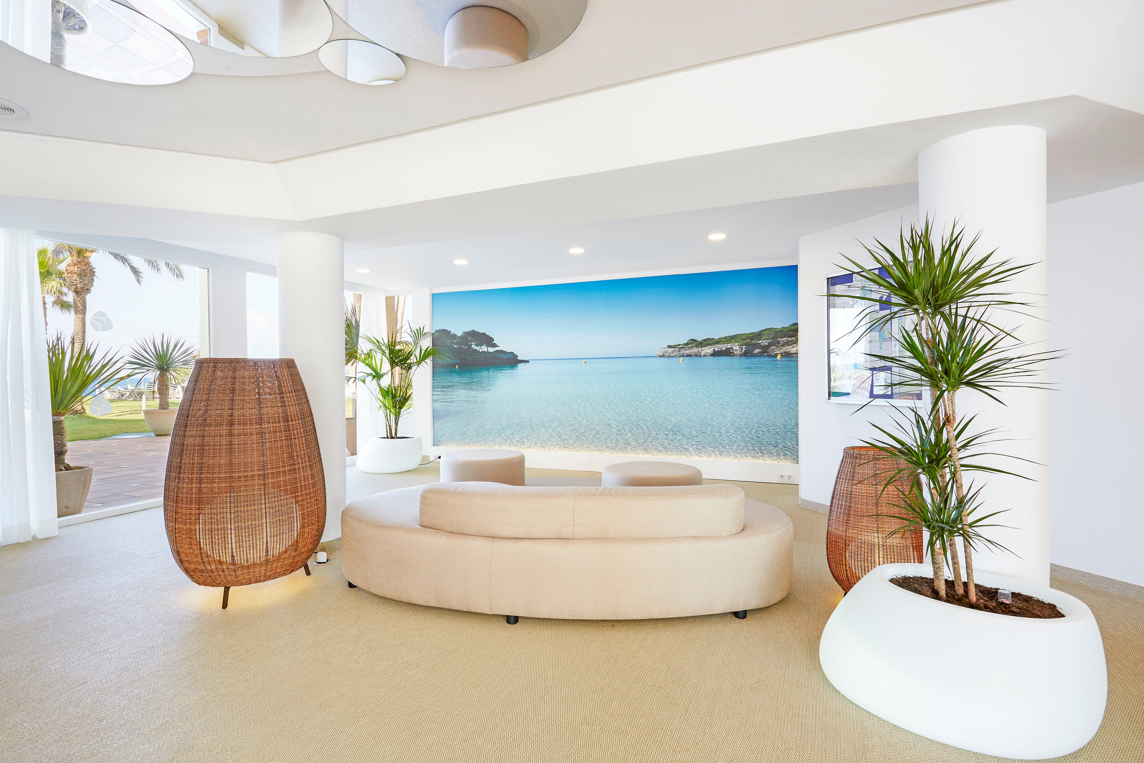 A contemporary lounge area features a curved beige sofa facing a large wall mural of a serene coastal bay. Tall white columns and expansive windows fill the space with natural light and views of palm trees outside. Woven rattan chairs and white planters with green indoor plants add organic texture to the minimalist interior. The overall atmosphere is airy, relaxed, and inspired by beachside living.