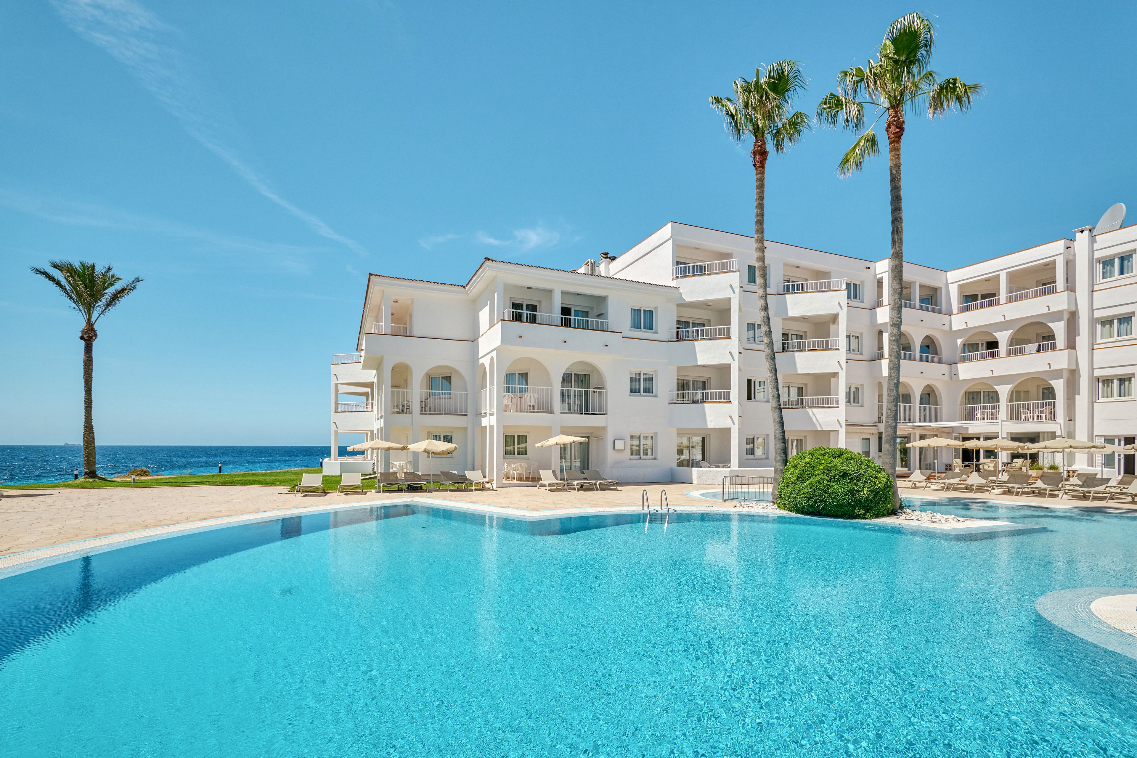 A bright seaside resort features a large turquoise swimming pool in the foreground. White multi-story buildings with balconies and arched windows stand behind the pool, facing the ocean. Tall palm trees and sun loungers with umbrellas line the poolside, creating a relaxing vacation atmosphere. The clear blue sky and calm sea enhance the sunny, tranquil coastal setting.