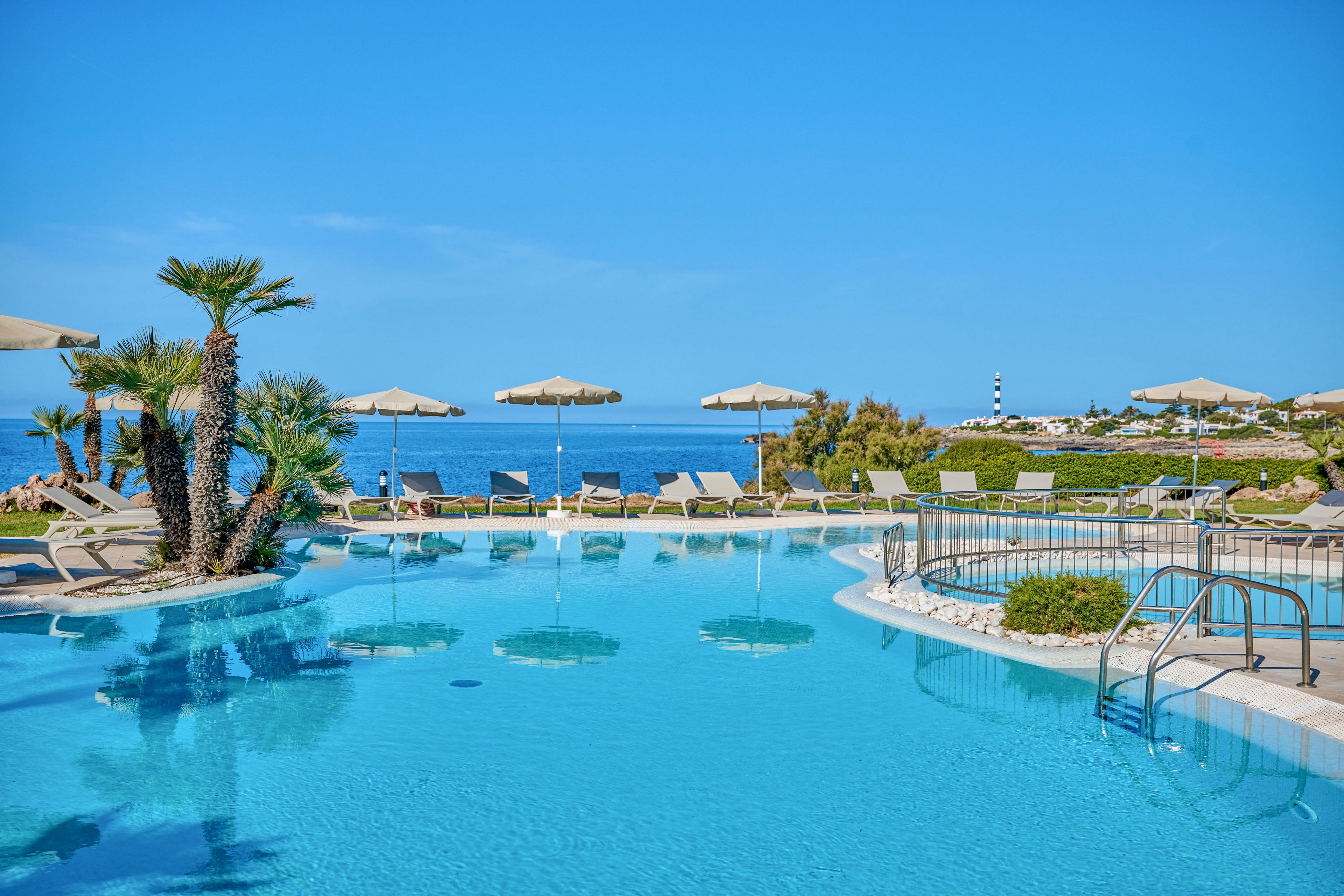 A clear blue swimming pool sits in the foreground of a coastal resort, surrounded by sun loungers and beige parasols. Palm trees and manicured greenery frame the pool area, leading the eye toward the calm sea in the background. A metal handrail and steps provide access to the water, while a distant lighthouse and low buildings dot the shoreline. The bright blue sky and vivid turquoise water create a relaxing, vacation atmosphere.