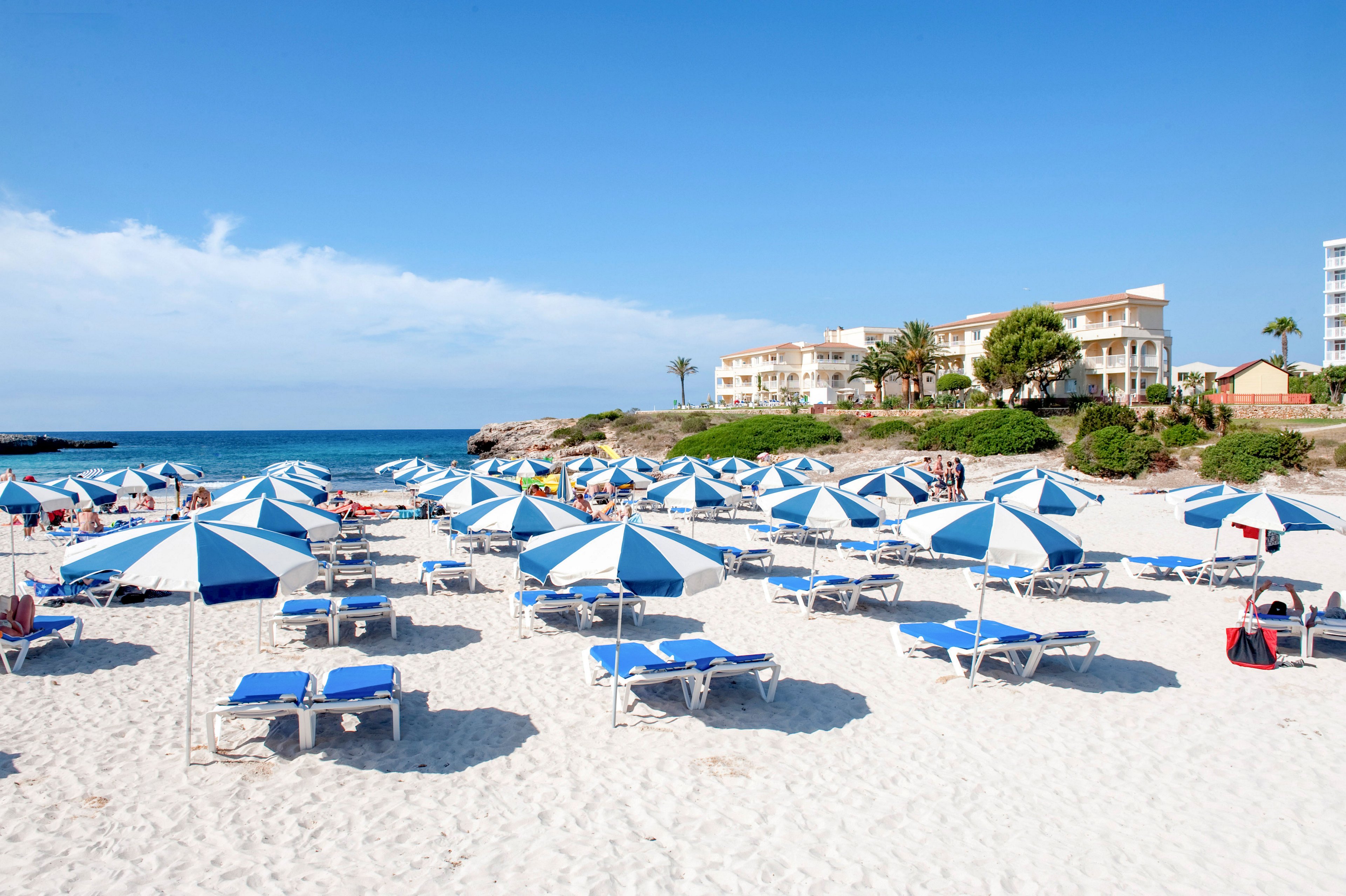 A bright sandy beach is lined with rows of blue and white umbrellas and matching sun loungers. The calm blue sea stretches into the distance under a clear sky. In the background, low-rise resort buildings and palm trees overlook the shoreline. A few people relax near the water, creating a peaceful holiday atmosphere.