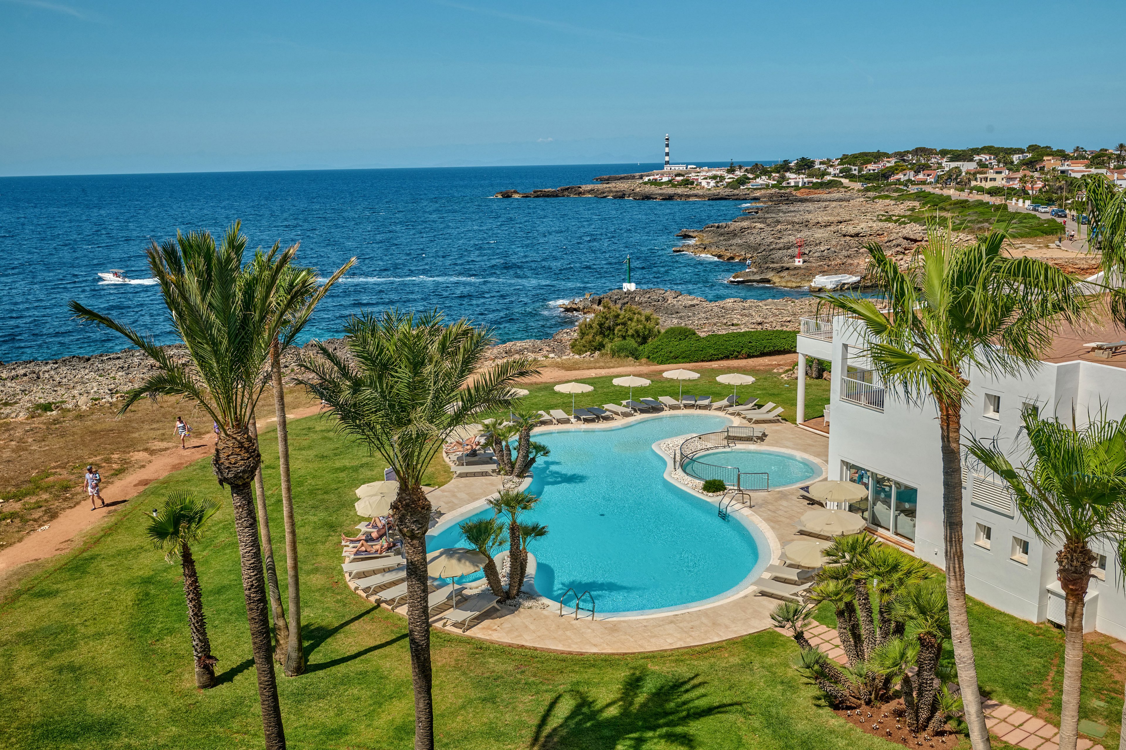 A curved swimming pool sits beside a white resort building, surrounded by tall palm trees and manicured green lawns. The property overlooks a rocky coastline with calm blue sea stretching into the distance. A lighthouse stands on a distant point, and a small boat moves across the water. Sun loungers and parasols around the pool suggest a relaxing holiday atmosphere in a sunny coastal destination.
