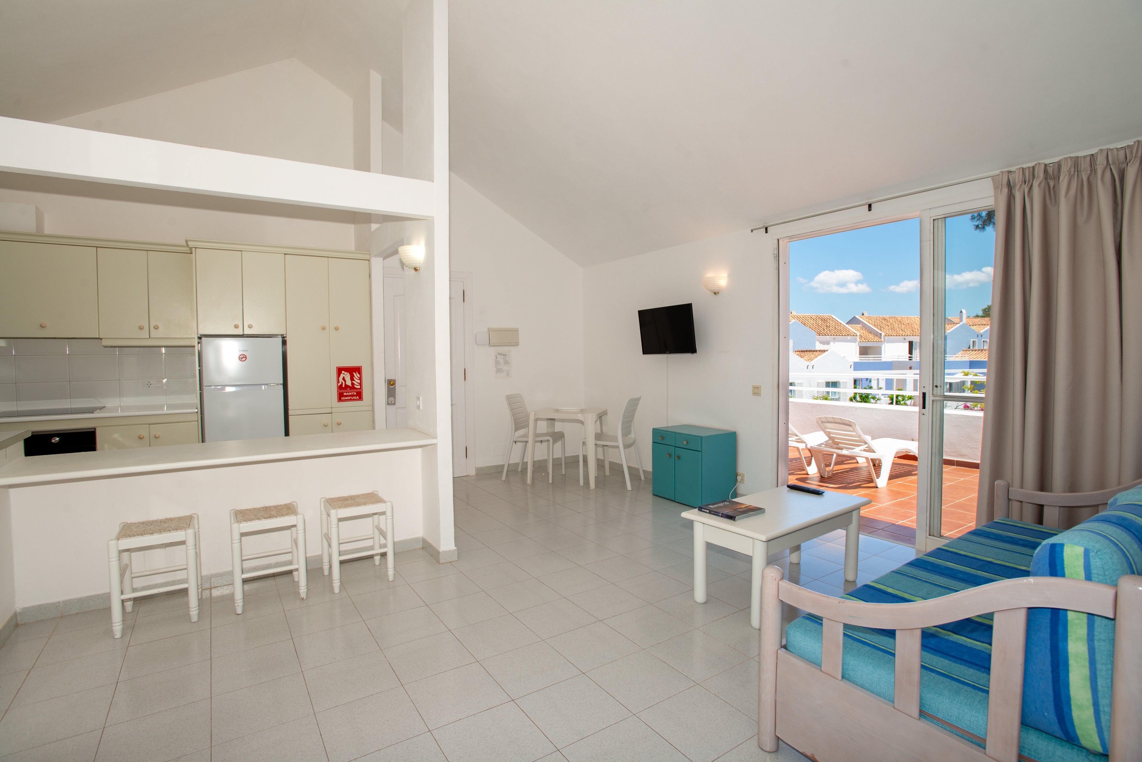 Spacious vacation apartment interior featuring an open-plan living room and kitchen. The room includes a tiled floor, white cabinetry, and a stainless steel refrigerator beside a breakfast bar with four stools. A striped sofa, wall-mounted television, and small table create a relaxed seating area. Large sliding doors open to a sunny balcony with outdoor chairs and views of neighboring houses under a blue sky.