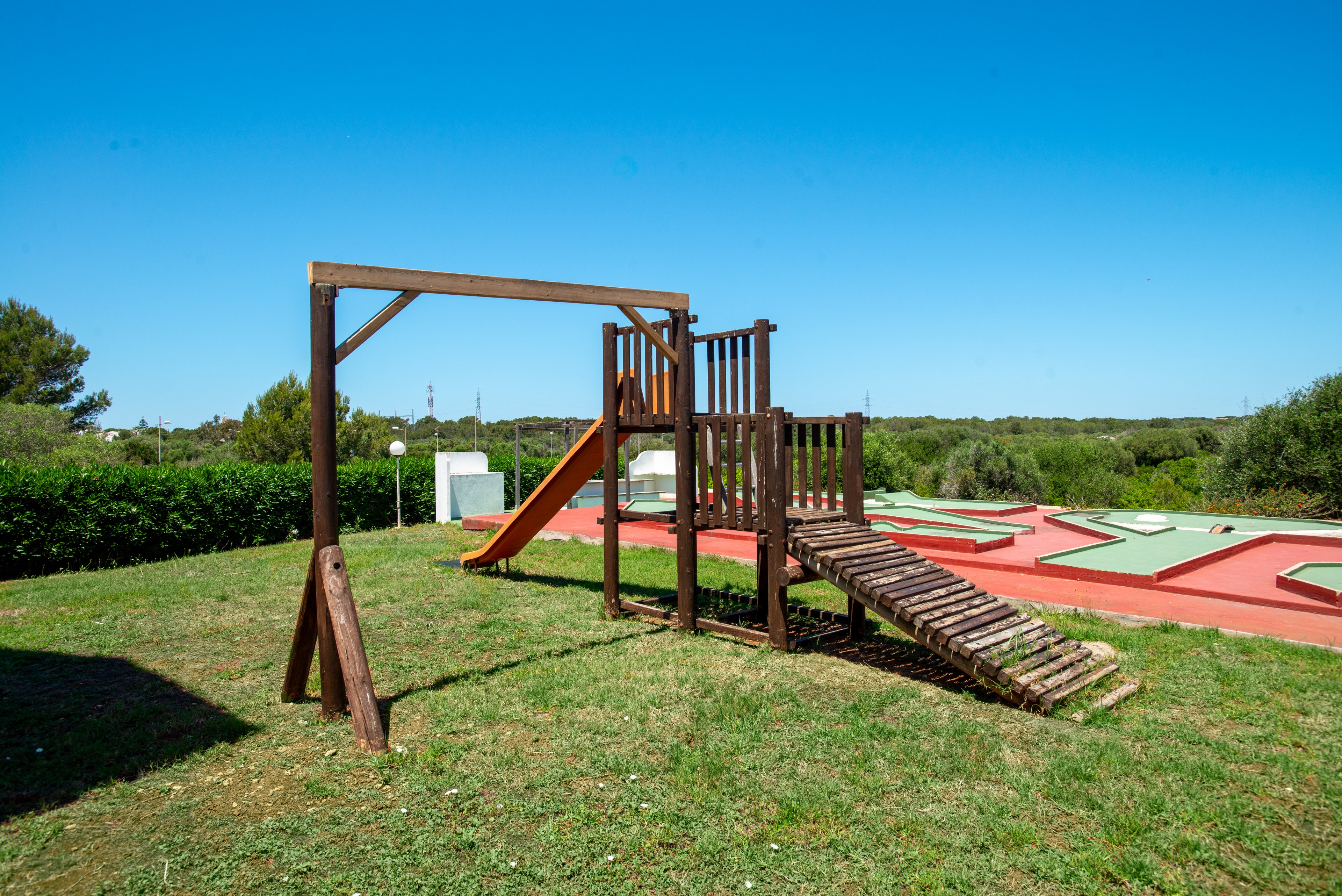 A wooden playground structure with a slide stands on a grassy lawn under a clear blue sky. The play equipment includes a small climbing bridge and support frame, set in an open outdoor park area. In the background, low hedges, trees, and a red-surfaced recreational zone are visible. The bright daylight and vivid colors create a cheerful, family-friendly atmosphere.