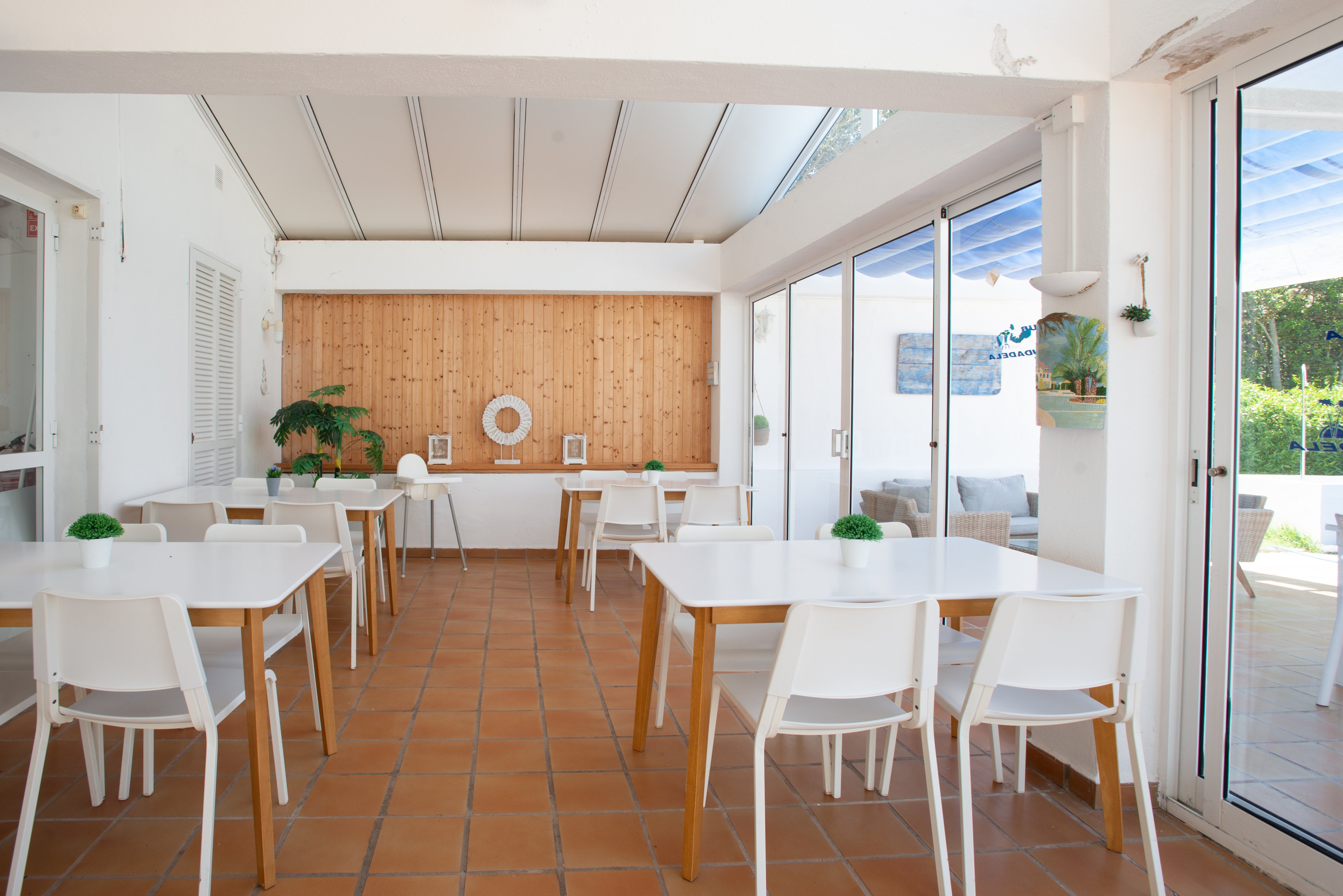 Clean white tables and chairs are arranged neatly in a bright, minimalist cafe interior. Large glass doors open to an outdoor terrace, allowing natural light to fill the space. Terracotta floor tiles contrast with white walls and a wooden accent wall featuring simple decor and plants. The overall atmosphere is airy, modern, and inviting, suitable for casual dining or coworking.