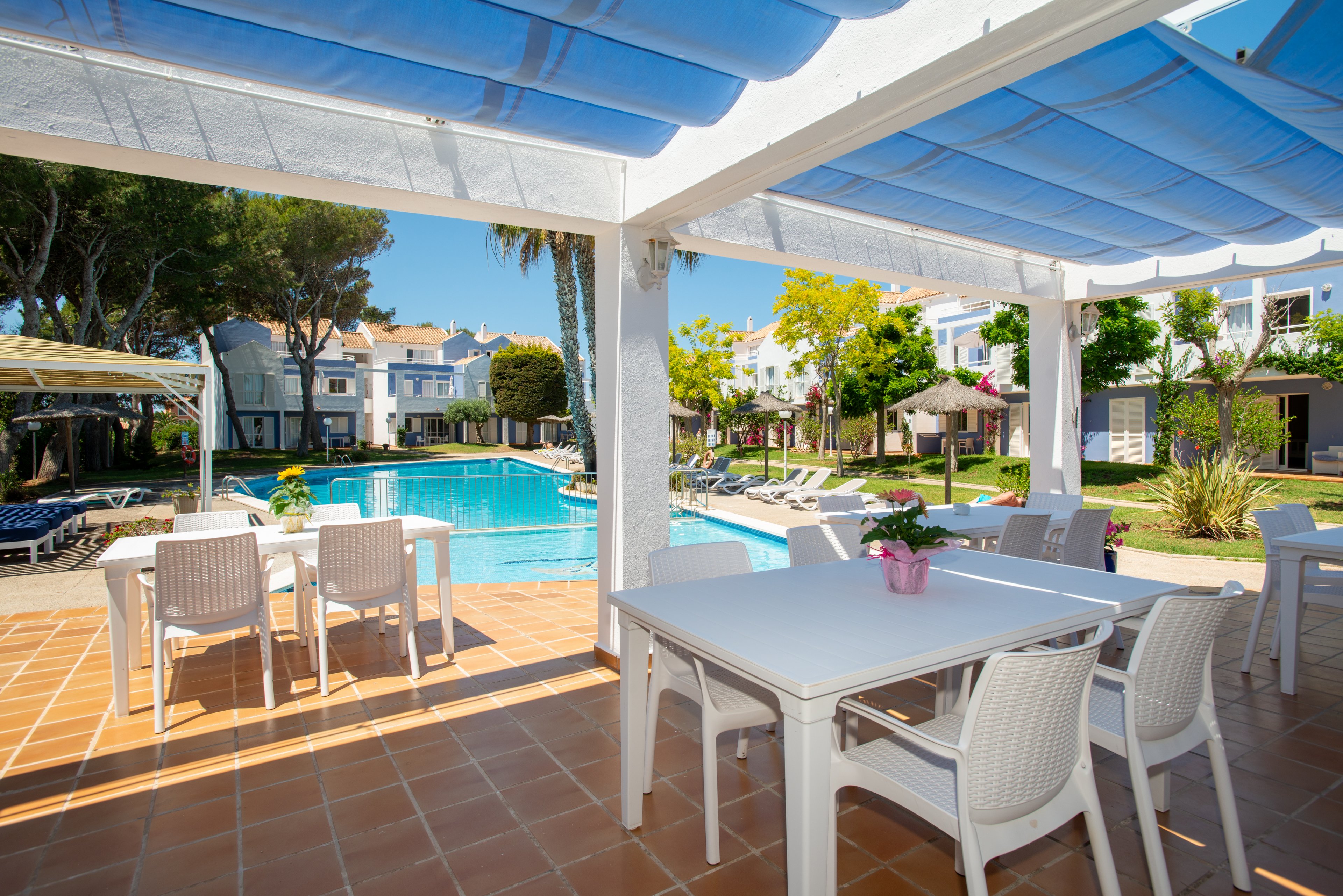 Bright outdoor dining tables sit under a shaded pergola beside a clear blue swimming pool. White lounge chairs and umbrellas line the pool deck, surrounded by green trees and landscaped gardens. In the background, white residential-style resort buildings with terracotta roofs create a relaxed vacation atmosphere. A small potted plant decorates the foreground table, adding a touch of color to the clean, modern setting.
