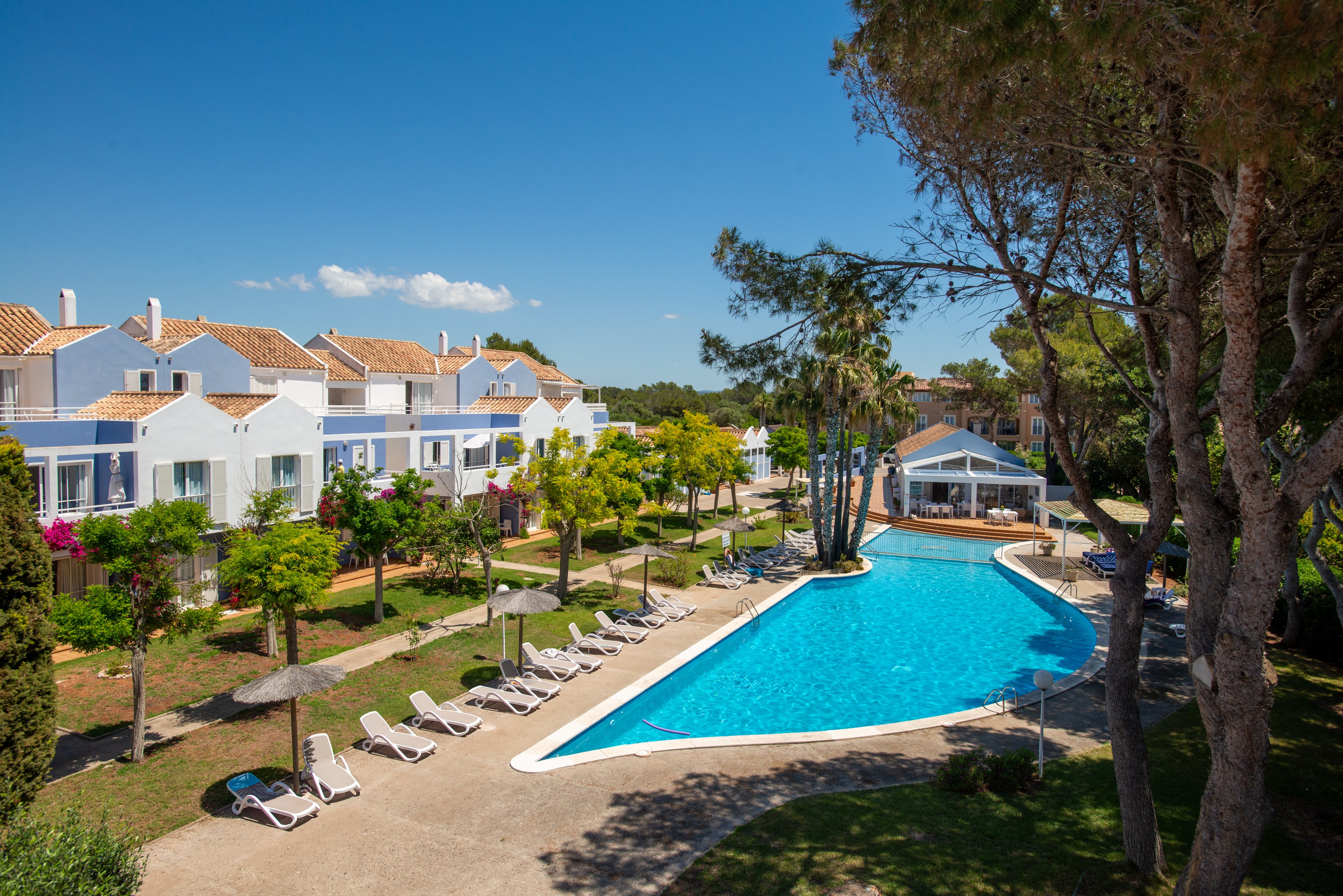 A long, bright blue swimming pool stretches through a landscaped resort lined with white and pastel-colored villas. Sun loungers and parasols are arranged neatly along the poolside, suggesting a relaxing holiday atmosphere. Tall trees and flowering shrubs add greenery and shade to the scene under a clear blue sky. The architecture and outdoor amenities evoke a peaceful Mediterranean-style vacation setting.