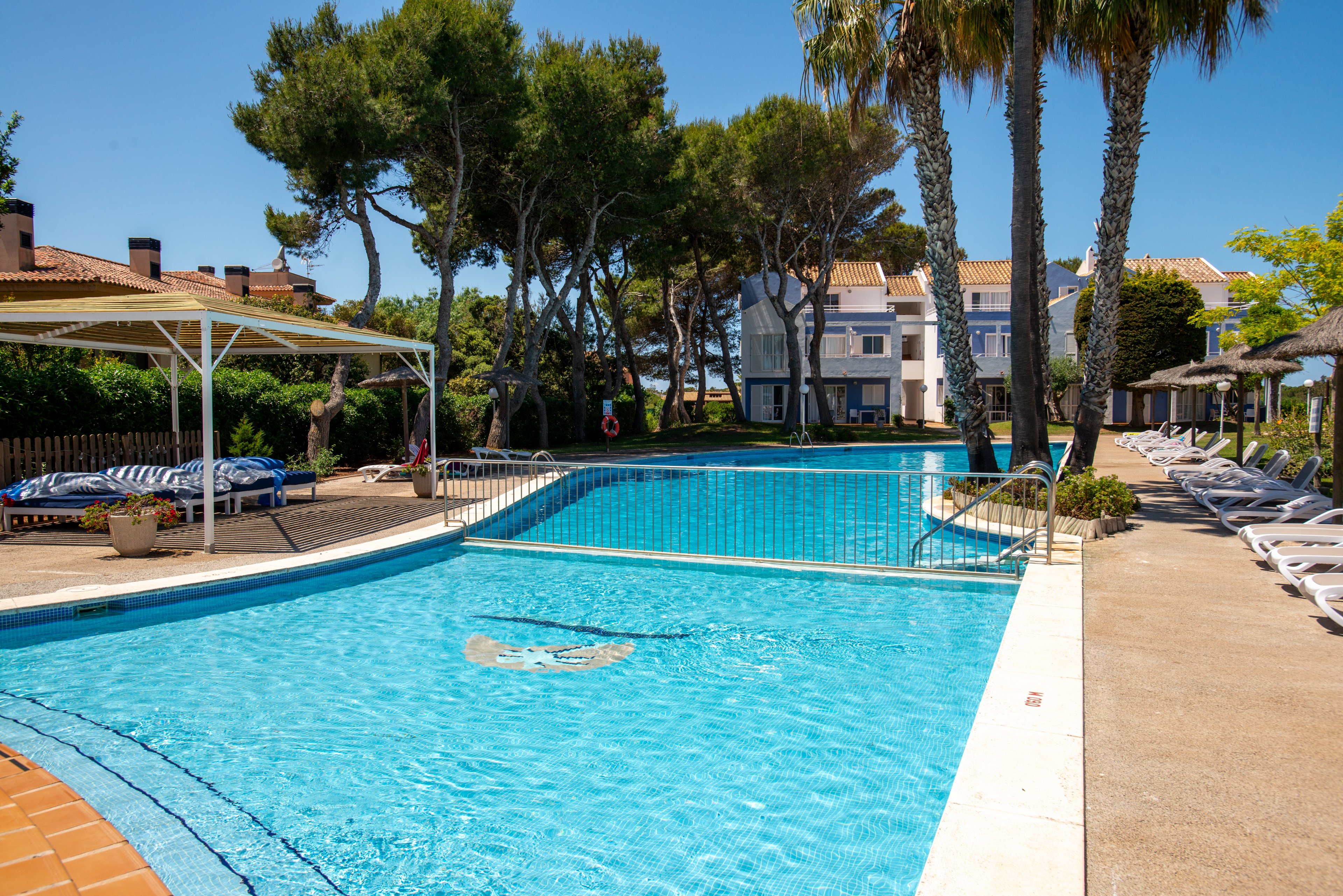 An outdoor swimming pool is shown on a bright sunny day at a resort or vacation property. Lounge chairs line the poolside, some shaded by umbrellas and a white pergola structure. Tall palm trees and dense green foliage surround the area, with residential-style buildings visible in the background. The clear blue water and relaxed seating create a tranquil holiday atmosphere.
