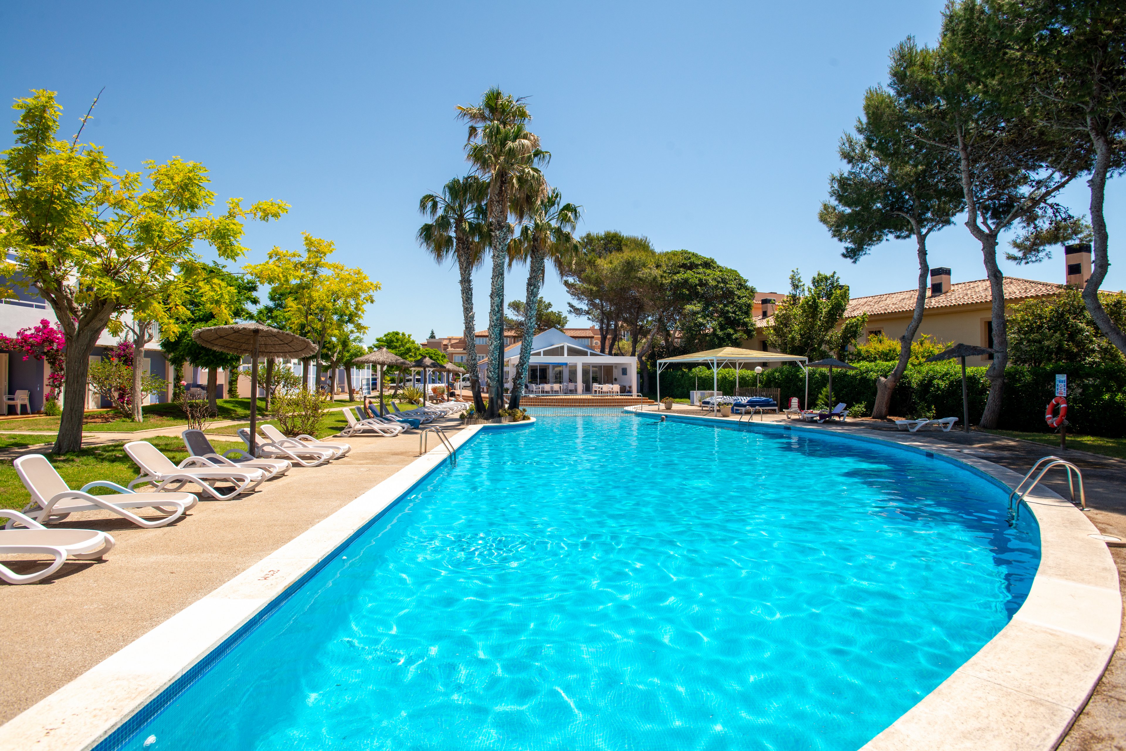 A bright blue outdoor swimming pool curves through a resort under a clear sky. White sun loungers line the poolside, shaded by umbrellas and surrounded by green trees. In the background, low-rise holiday buildings and palm trees create a relaxed vacation atmosphere. The scene suggests a warm, sunny day ideal for leisure and relaxation.
