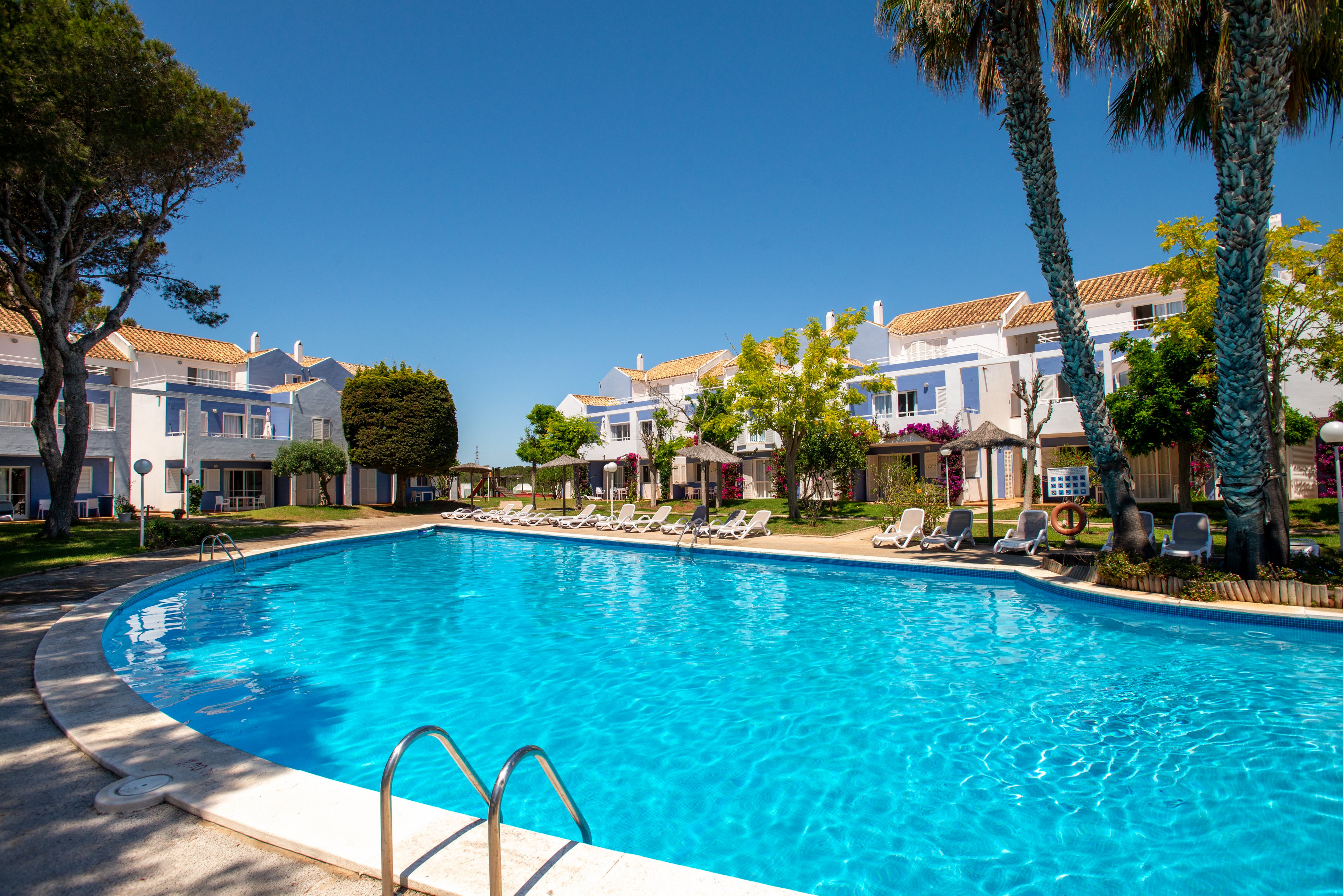 Large outdoor swimming pool at a resort is surrounded by white two-story buildings under a clear blue sky. Sun loungers and umbrellas line the poolside, suggesting a relaxing vacation setting. Tall trees and manicured greenery add a touch of nature to the bright, sunny scene. The calm water and empty chairs create a peaceful, inviting atmosphere.