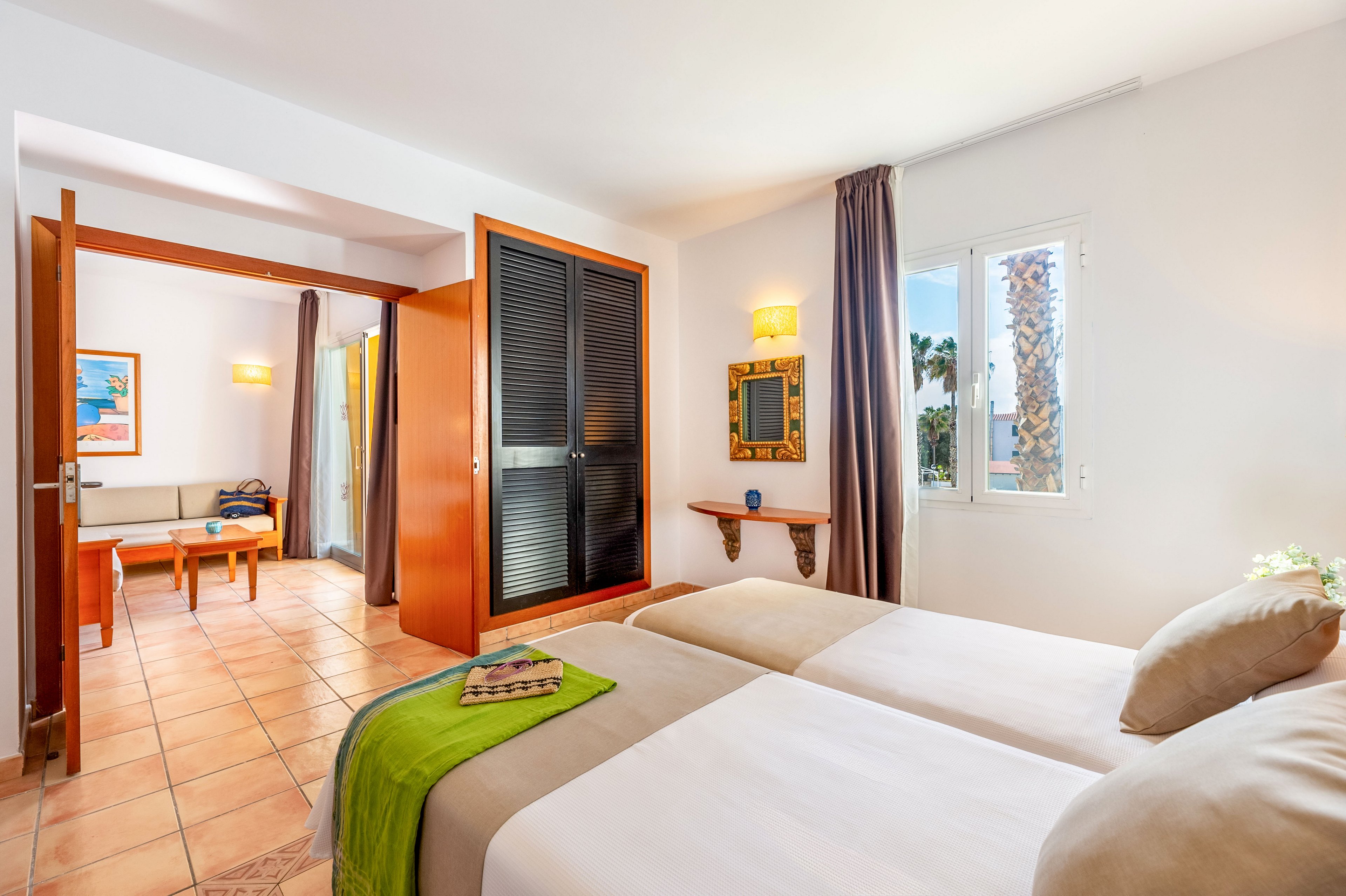 Well-lit hotel room featuring two neatly made twin beds with neutral bedding and a green throw. A small window lets in natural light and offers a view of palm trees outside. Warm-toned tile flooring, wooden furniture, and soft wall lighting create a welcoming atmosphere. An open doorway leads to an adjoining sitting area with a table and chairs, enhancing the spacious feel of the accommodation.