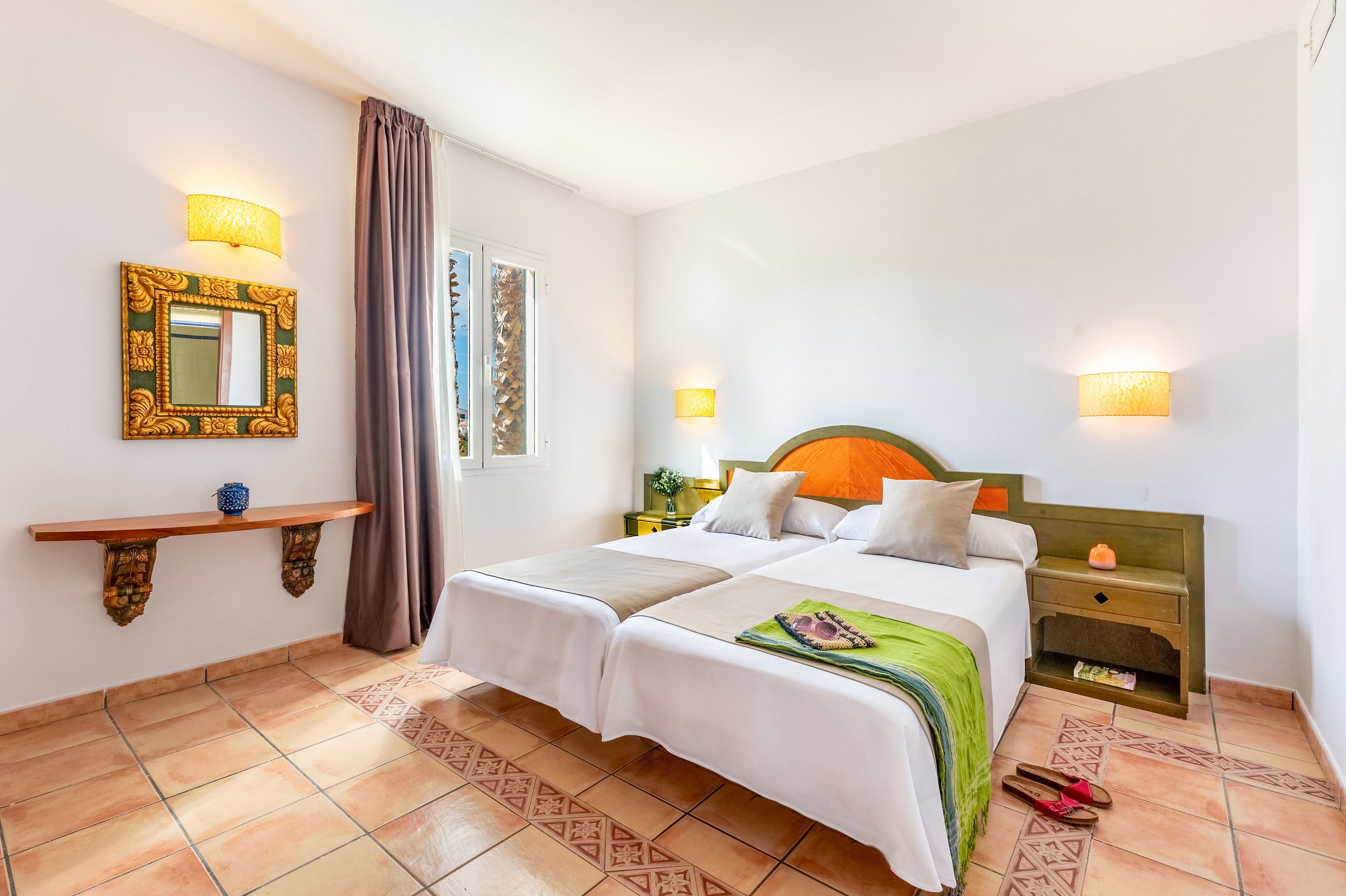 Sunny hotel room featuring two neatly made twin beds with white linens and green runners. The space includes a tiled floor, bedside table with lamp, and a decorative wall mirror above a wooden shelf. A window with long curtains lets in natural light, highlighting a magazine and sandals placed casually near the bed. Warm wall sconces and soft colors create a welcoming, relaxed atmosphere.