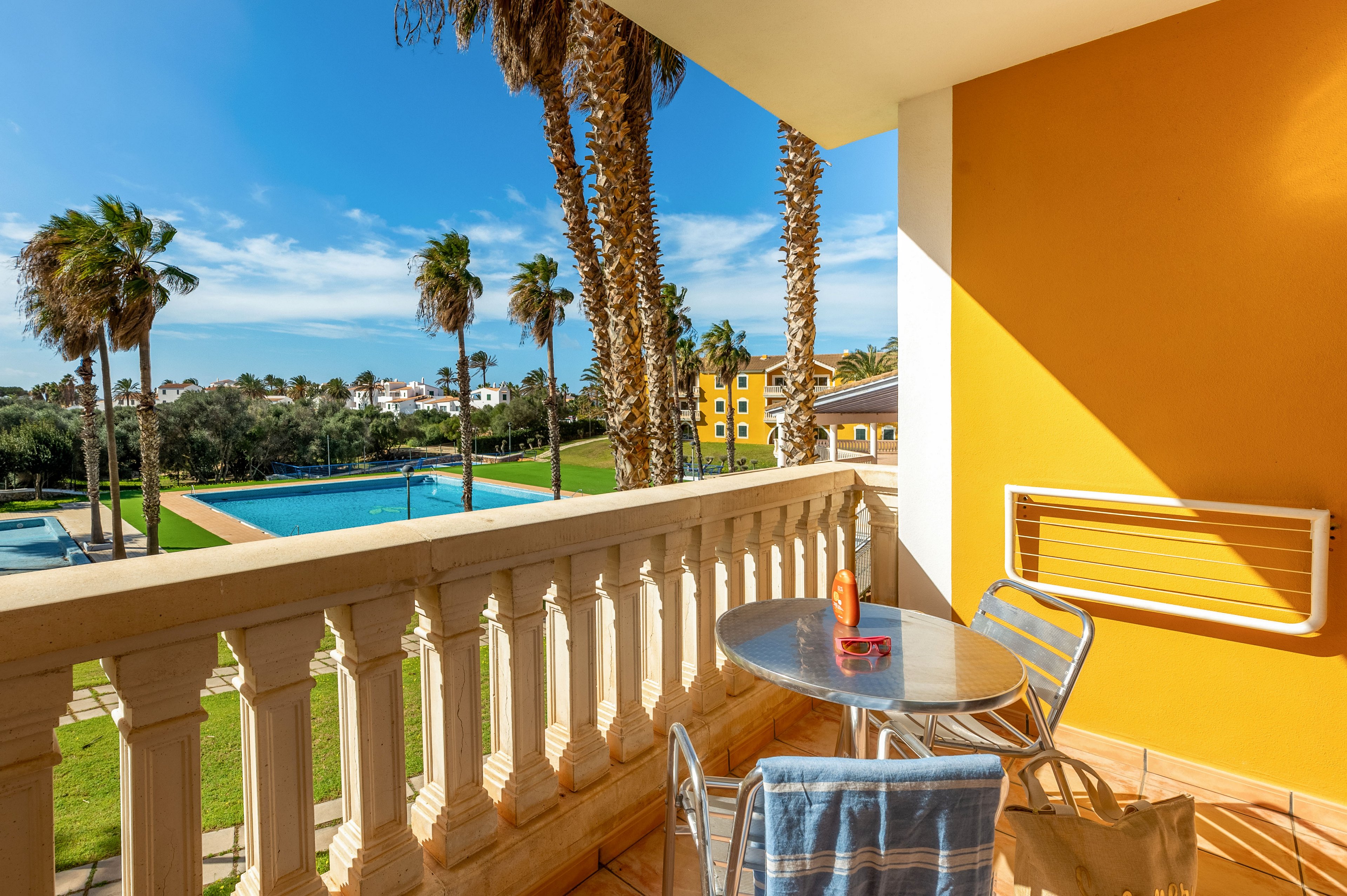 A bright balcony with a small round table and two chairs overlooks a landscaped pool area and the distant ocean. A drink bottle and glass sit on the table, while a striped towel hangs over the balcony railing. Tall palm trees and green lawns lead toward colorful resort buildings under a clear blue sky. The warm yellow wall and tiled floor create a vibrant, inviting holiday atmosphere.