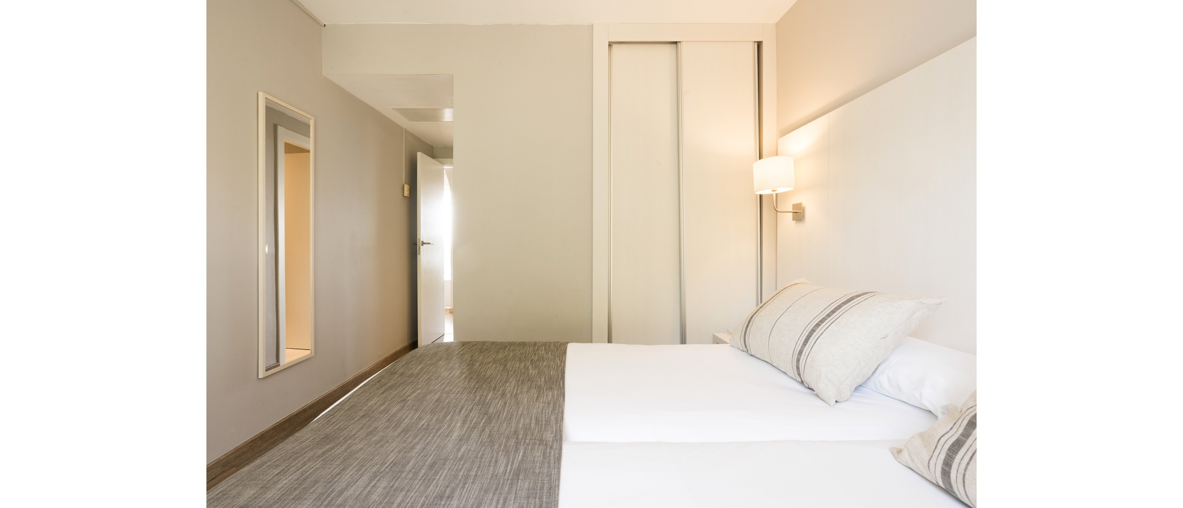 A bright, minimalist bedroom features a neatly made double bed with neutral-toned bedding. The room has light-colored walls, a wall-mounted lamp, and a large mirror reflecting part of the space. Sliding closet doors and an open doorway add depth and structure to the composition. Soft natural light enhances the clean, modern aesthetic of the interior.