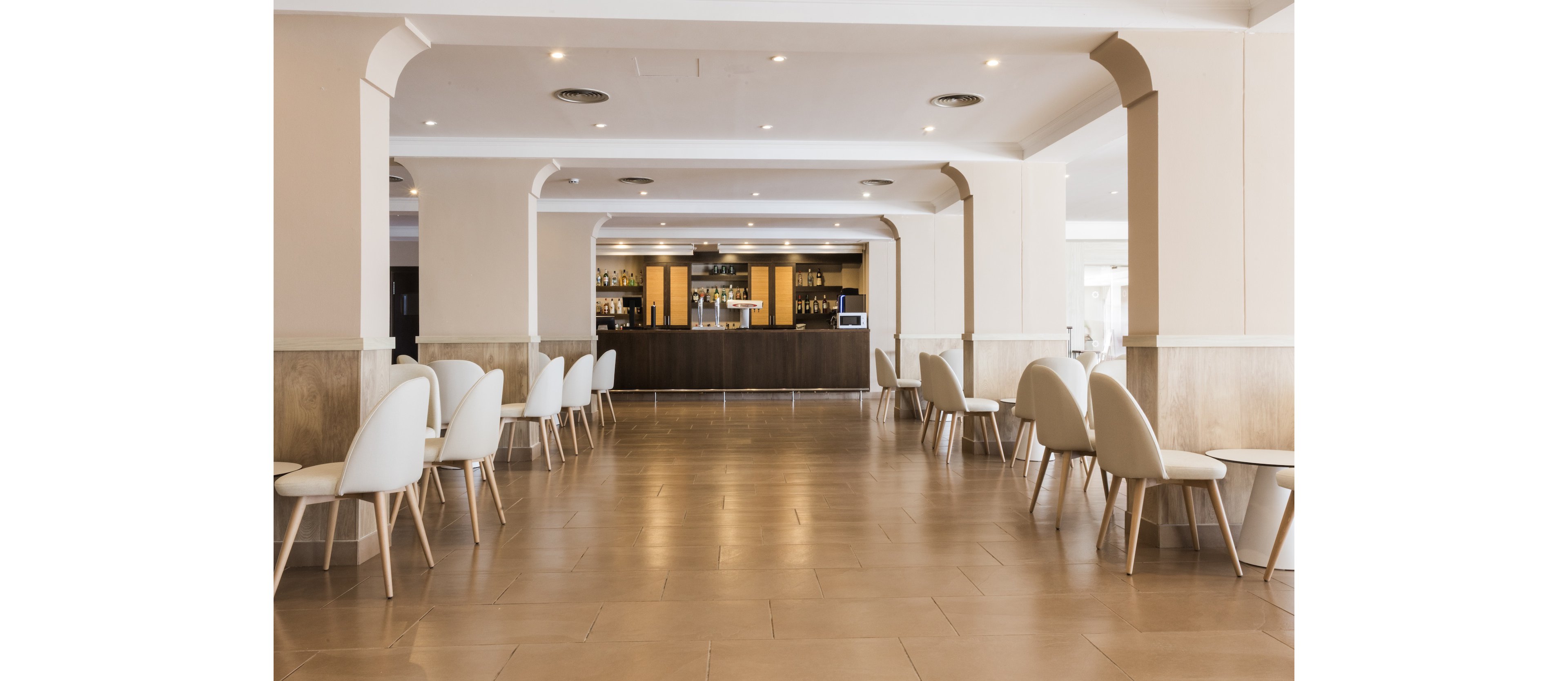 A spacious hotel lobby features a long corridor leading to a bar at the far end. White contemporary chairs and small round tables are arranged symmetrically along both sides of the polished floor. Soft neutral tones and clean architectural lines create a minimalist, modern atmosphere. Recessed ceiling lights and natural light enhance the bright, welcoming interior.