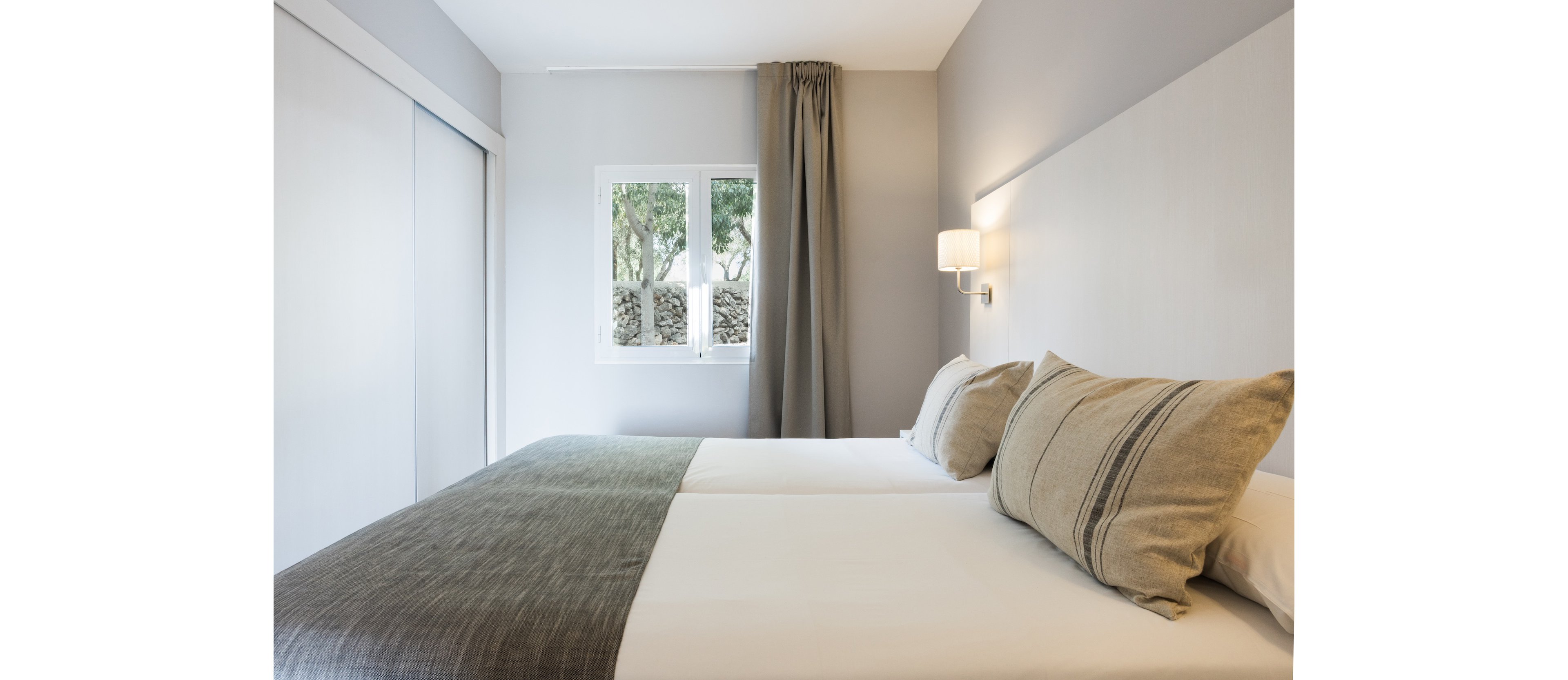 A bright, minimalist bedroom features two neatly made twin beds with neutral-toned linens and accent pillows. A small window lets in natural light and offers a view of trees and a stone wall outside. Soft gray curtains and walls create a calm, contemporary atmosphere. A wall-mounted lamp adds warm lighting to the serene interior.