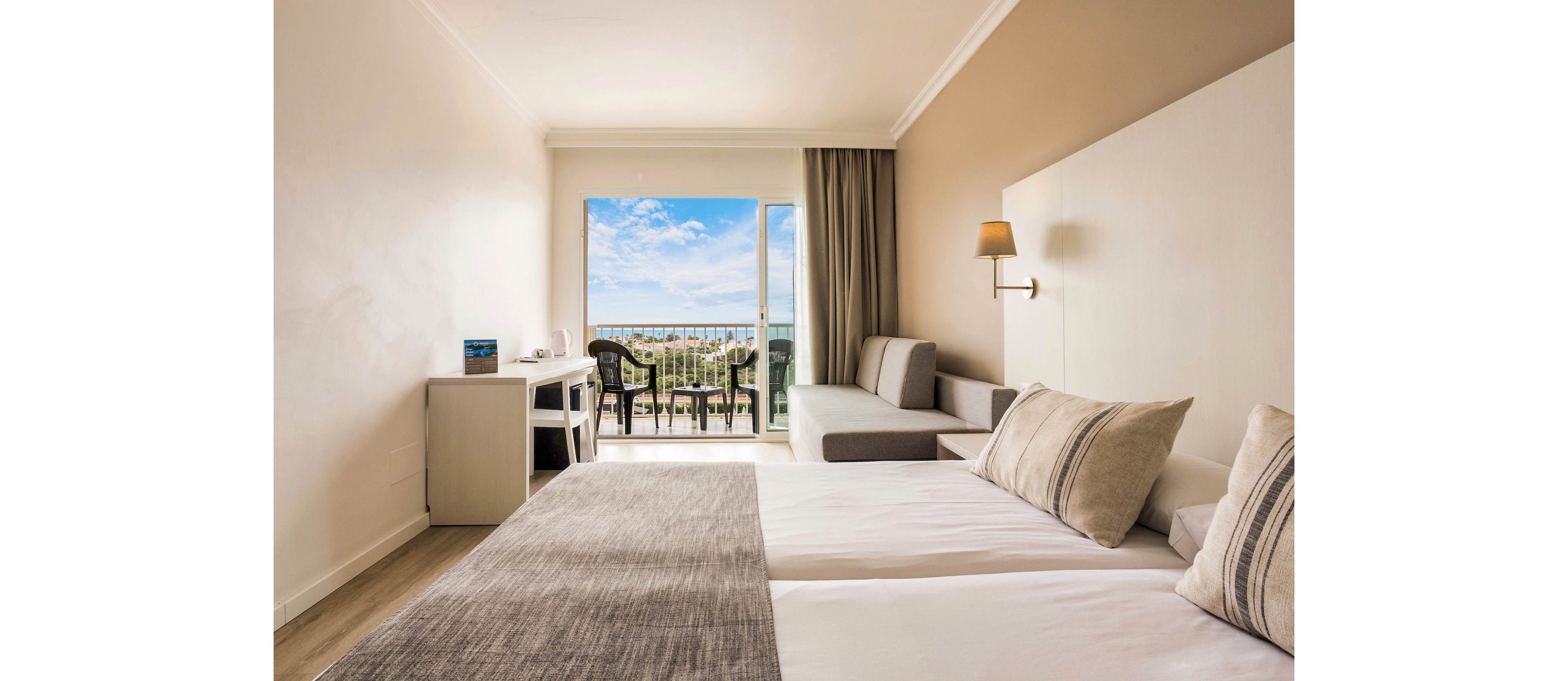 A modern hotel room features two neatly made beds with neutral-toned linens and cushions. The space includes a small desk, chair, and a gray sofa along the wall, all in a minimalist style. Large sliding glass doors open onto a balcony with black chairs and a table, overlooking greenery and a distant sea under a blue sky with clouds. Soft natural light fills the room, creating a calm and inviting atmosphere.
