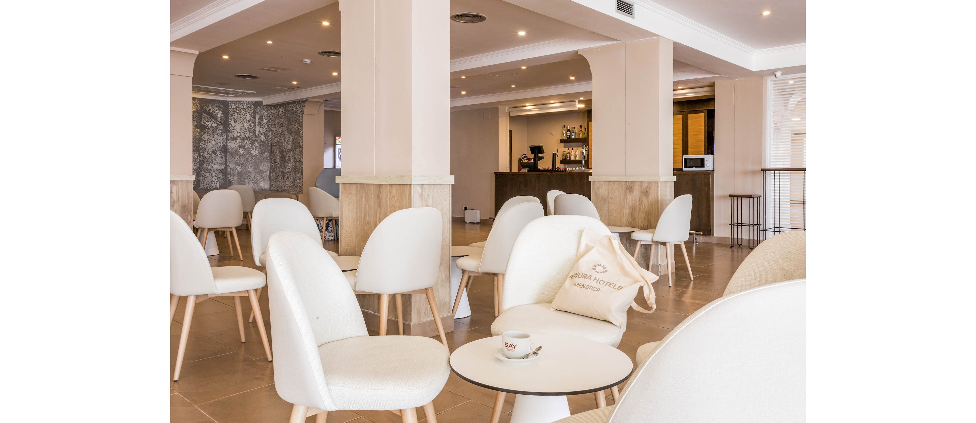 A bright, modern hotel lounge features rows of white chairs and small round tables arranged neatly across a spacious room. Warm neutral tones on the walls and floor create a calm, minimalist atmosphere. A coffee bar with bottles and glassware is visible in the background, adding a casual café feel. A tote bag and a cup on the foreground table suggest a relaxed, welcoming space for guests to sit and unwind.