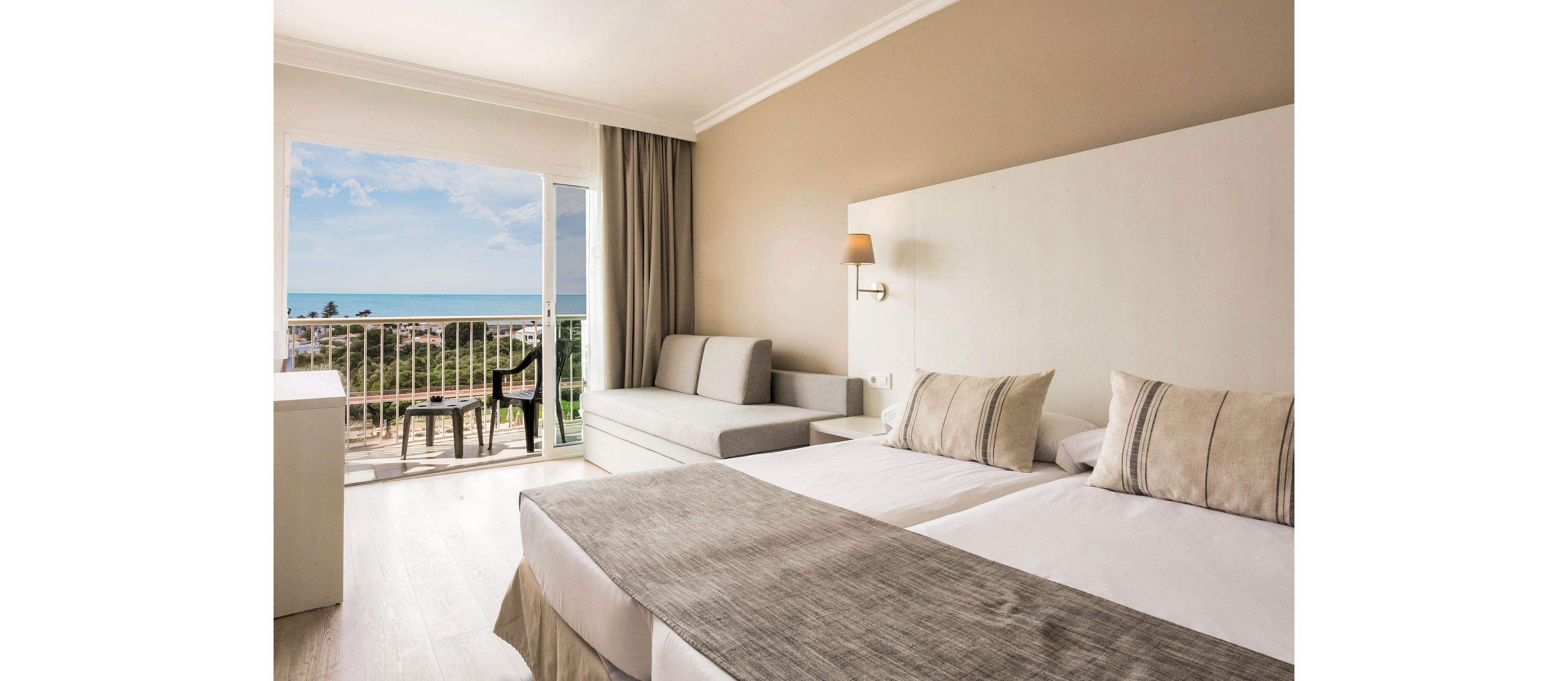 A modern hotel room features two neatly made twin beds with neutral-toned linens and accent pillows. A light gray sofa sits against the wall beside a small wall-mounted lamp, creating a cozy seating area. Large sliding glass doors open to a balcony with chairs and a table, overlooking trees and a distant ocean under a blue sky. Soft beige and white walls, sheer curtains, and natural daylight give the space a calm, airy atmosphere.