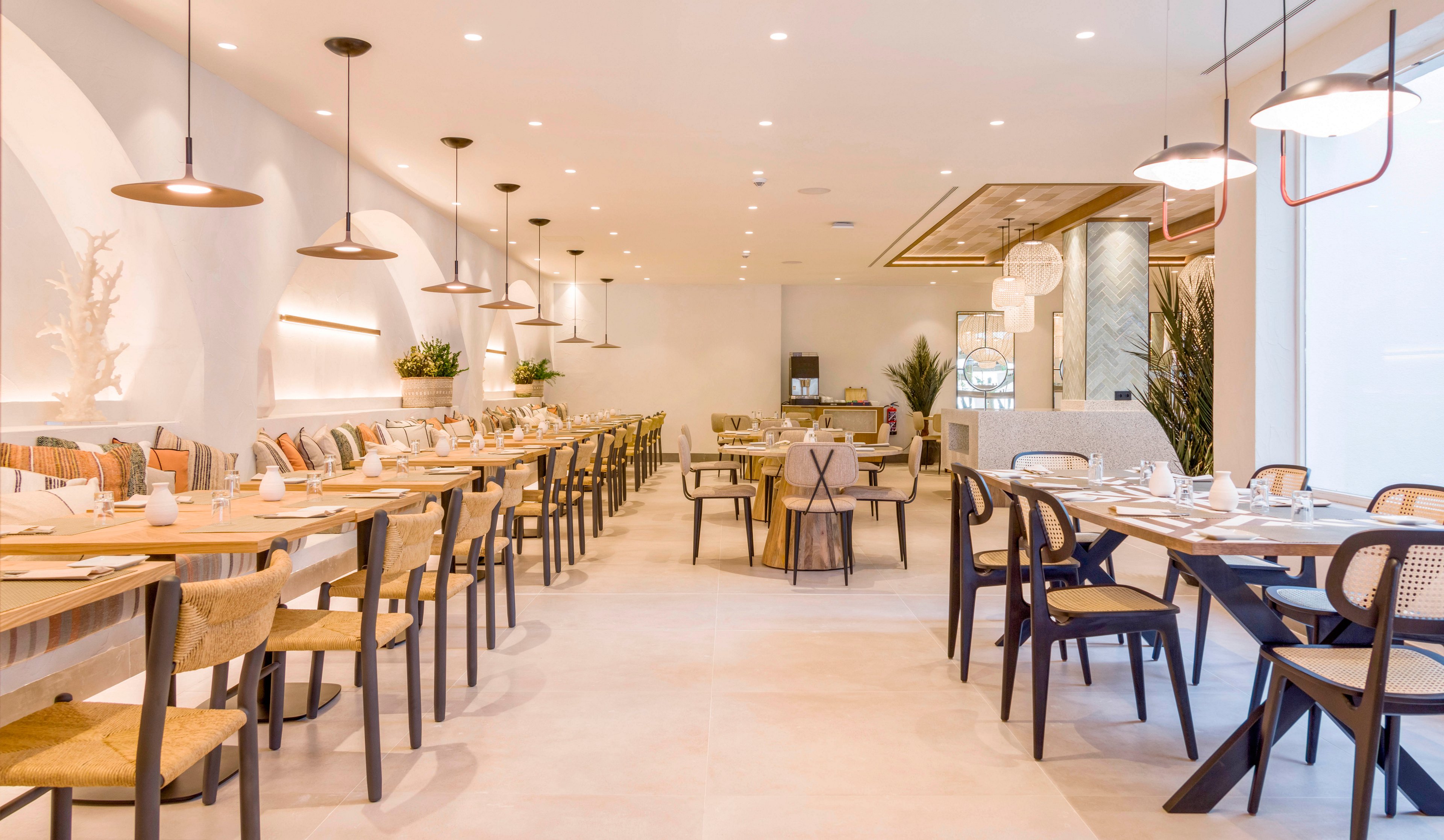Contemporary restaurant interior featuring long wooden tables neatly set with plates, cups, and cutlery. The space is bright and airy with white walls, light flooring, and warm pendant lighting. Mixed wooden and black chairs line both sides of the room, creating a clean and minimalist dining atmosphere. Green plants and subtle decorative elements add a touch of nature and softness to the modern design.