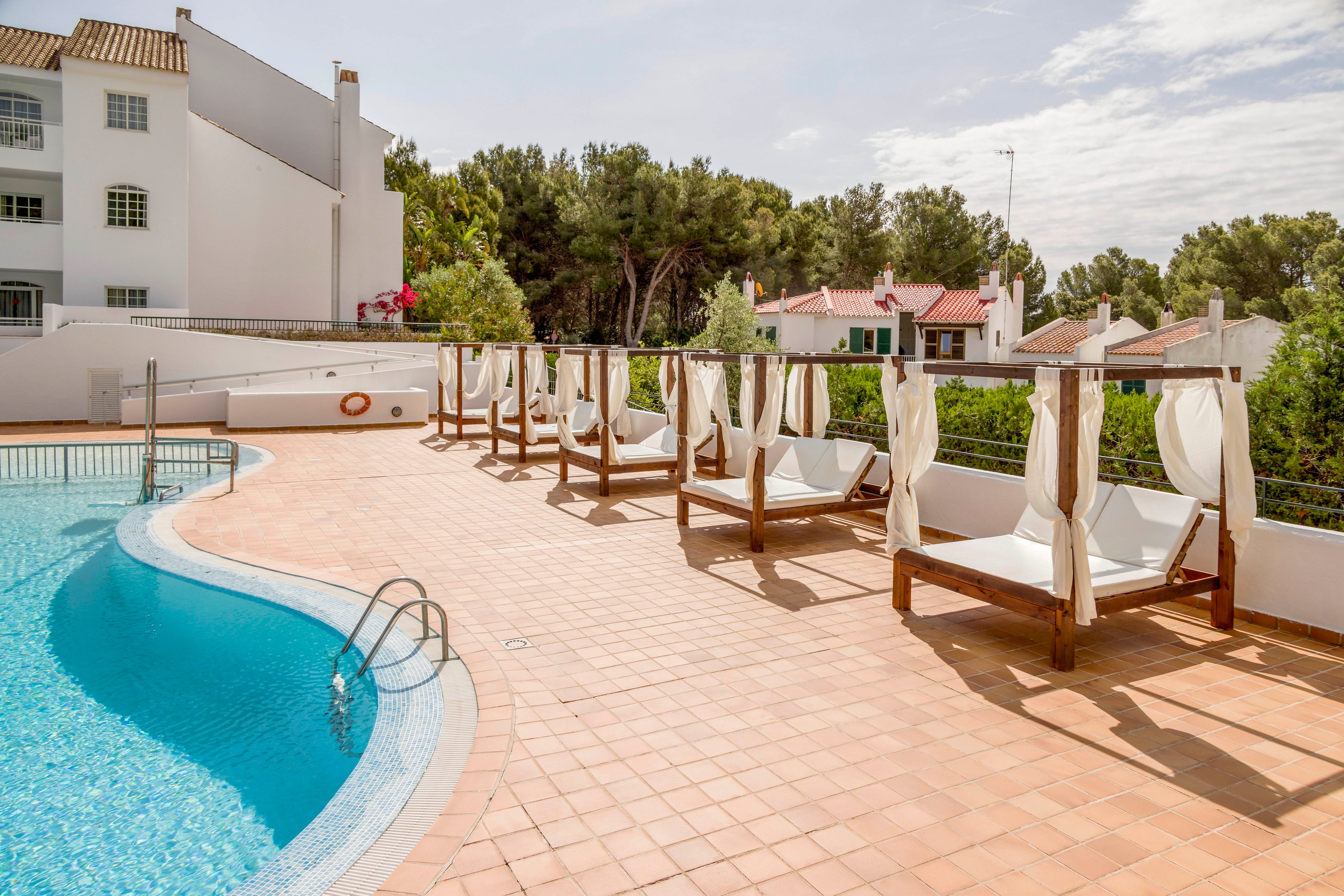 Outdoor swimming pool area with curved blue water is lined with wooden cabanas and white cushioned loungers. The scene is set at a quiet resort or apartment complex with white buildings and terracotta-tiled pool deck. Green trees and neighboring houses appear in the background under a bright, partly cloudy sky. The overall atmosphere is calm, inviting, and ideal for relaxing by the water.