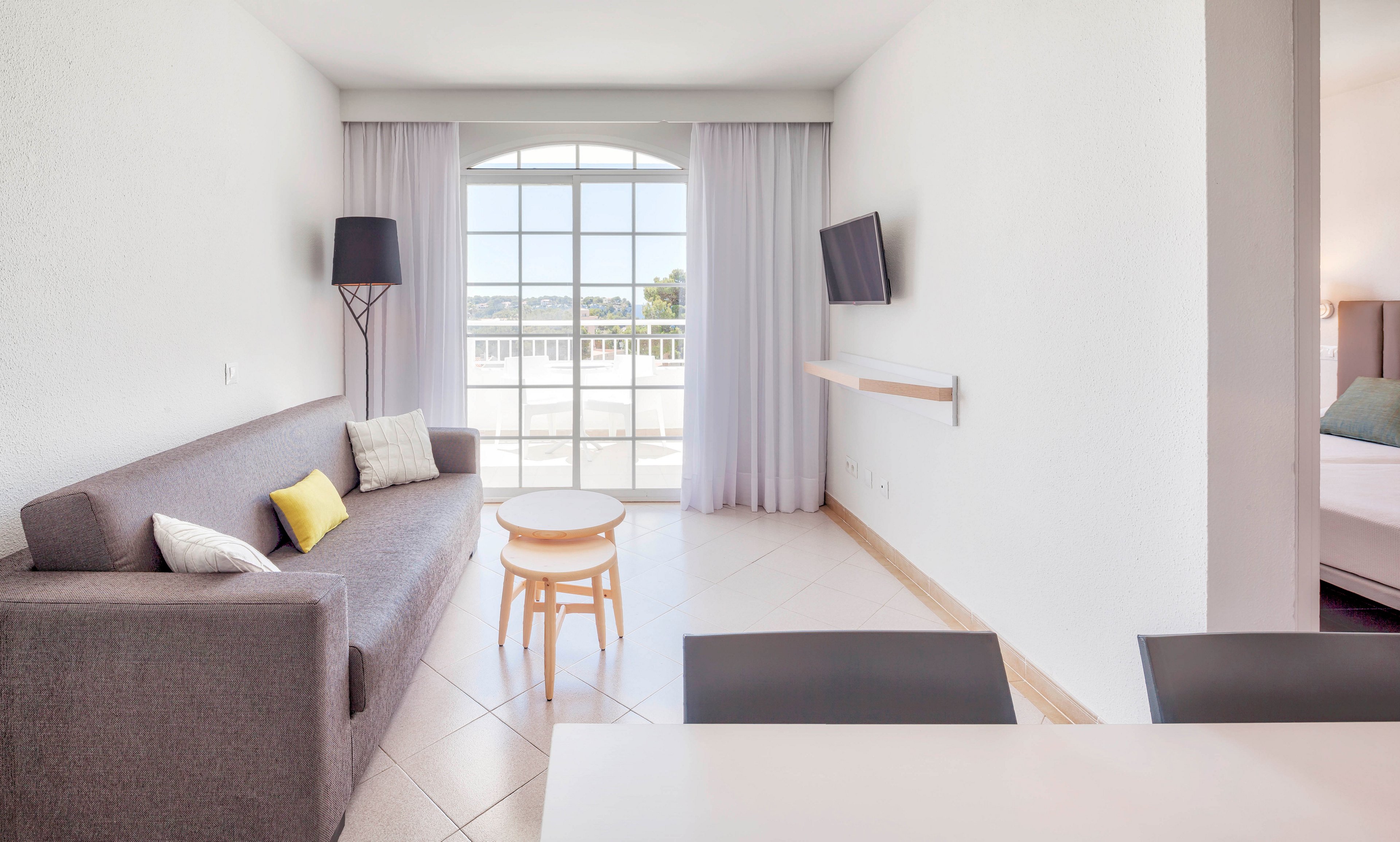 A bright, minimalist living room features a gray sofa with colorful accent pillows and a small round nesting table. Large glass doors with sheer white curtains open to a sunny balcony and distant landscape view. A wall-mounted flat-screen TV and modern floor lamp add contemporary touches to the clean white interior. The adjoining bedroom is partially visible, enhancing the sense of open-plan space and vacation comfort.