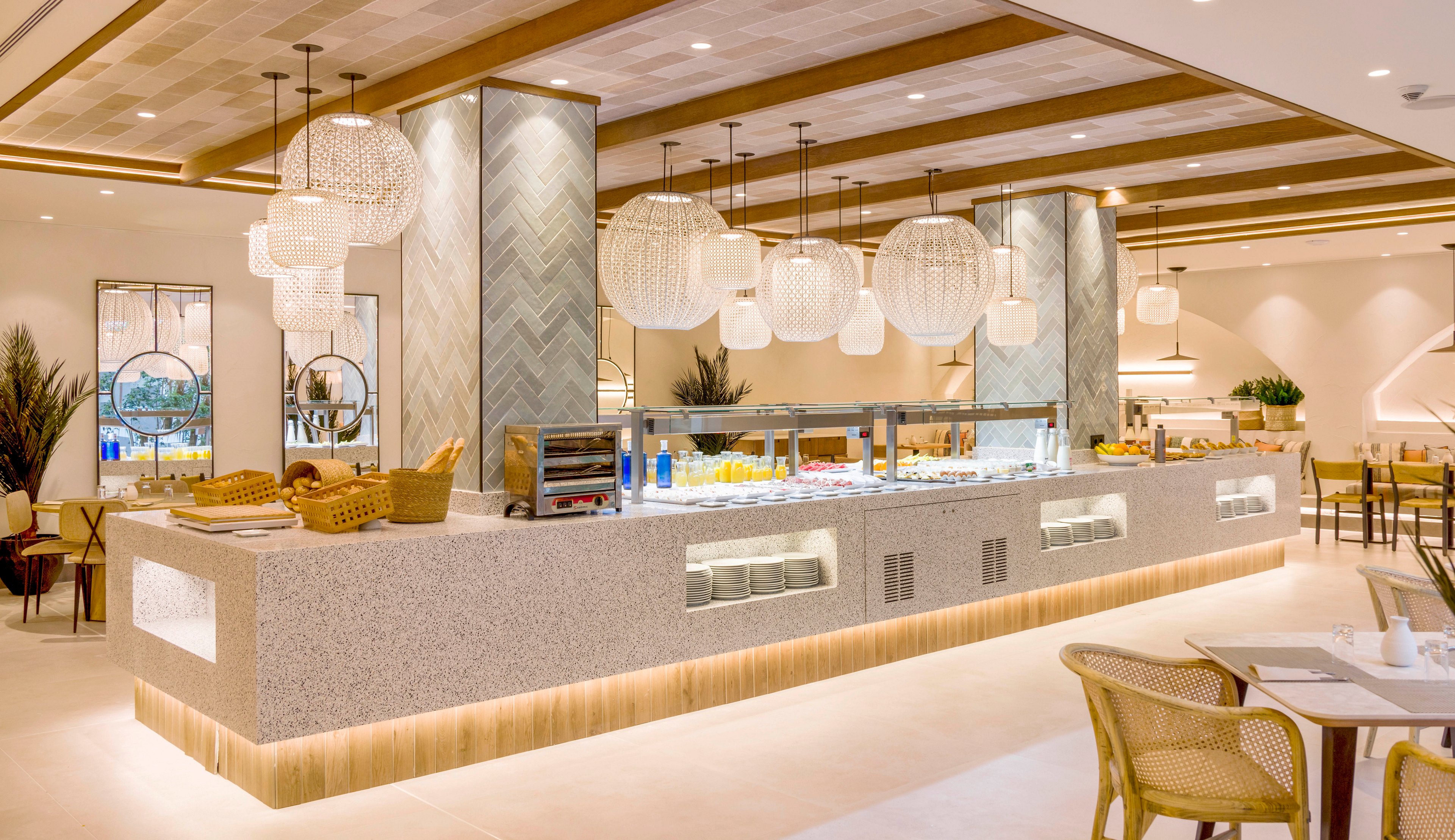 A spacious self-service buffet counter stands in the center of a modern restaurant. The setting features warm neutral tones, wooden ceiling beams, and large decorative pendant lights. Food trays and baskets are arranged neatly along the stone countertop, with seating areas visible in the background. The overall atmosphere is clean, contemporary, and inviting, suitable for hotel dining or resort breakfast service.