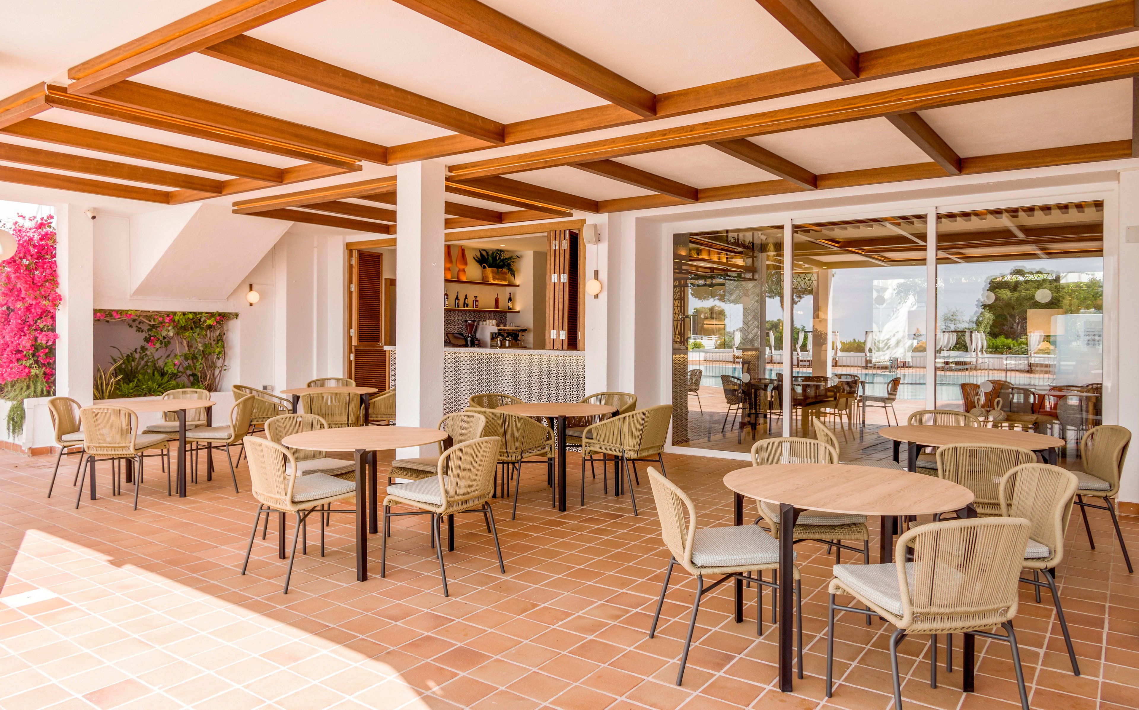 Open-air terrace café with several round tables and wicker chairs is arranged on a terracotta tiled floor. A small bar with bottles and glasses is visible in the background under a wooden beamed ceiling. Large sliding glass doors reveal a bright poolside area and greenery outside. Pink flowering plants add a touch of color to the otherwise neutral, modern resort setting.