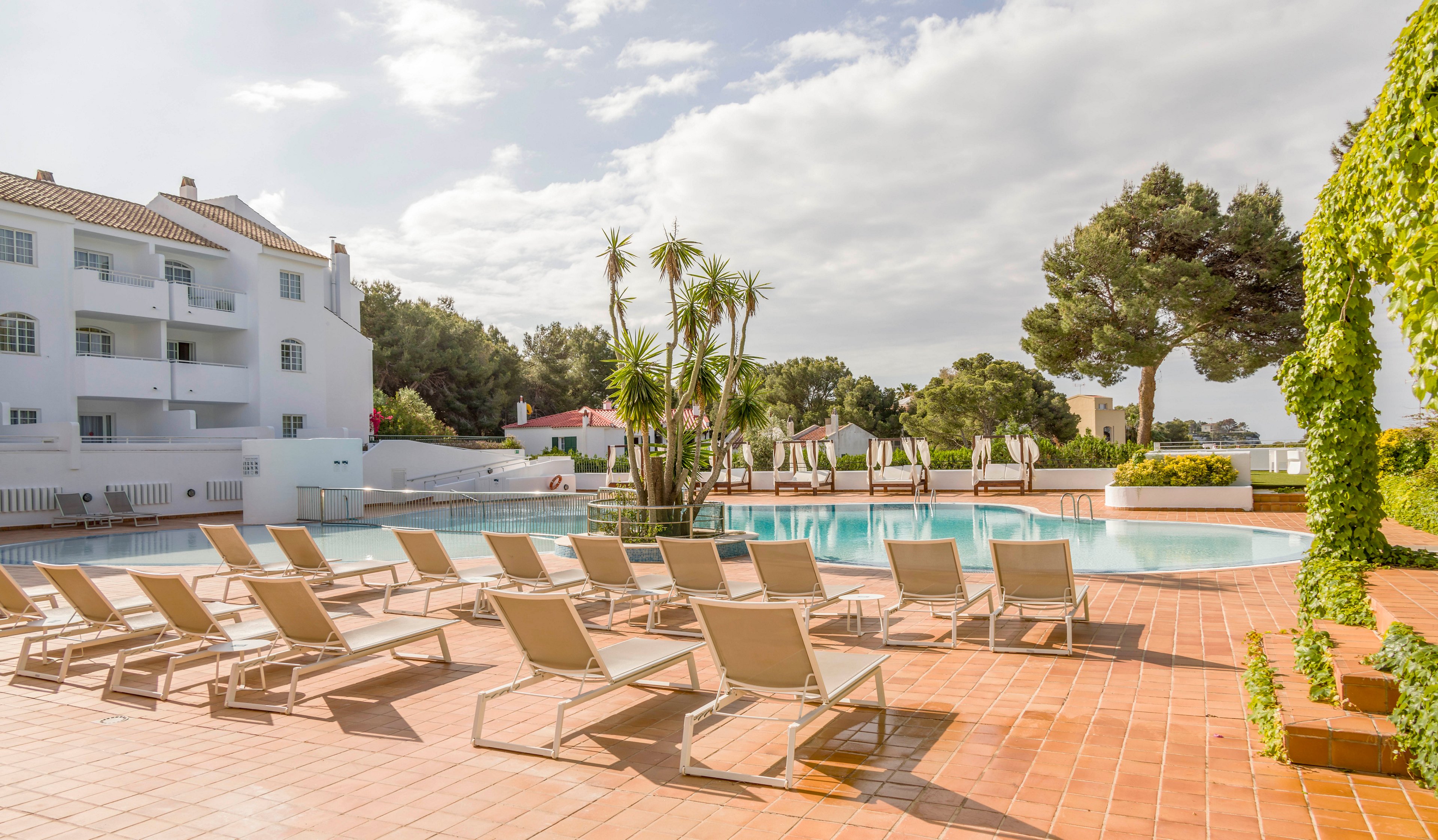 An outdoor swimming pool is surrounded by rows of empty sun loungers on a sunny day. The scene is set in a resort or apartment complex with white buildings and balconies in the background. Tall palm trees and lush greenery add a tropical feel around the pool area. The overall atmosphere is calm and inviting, suggesting a relaxing vacation destination.