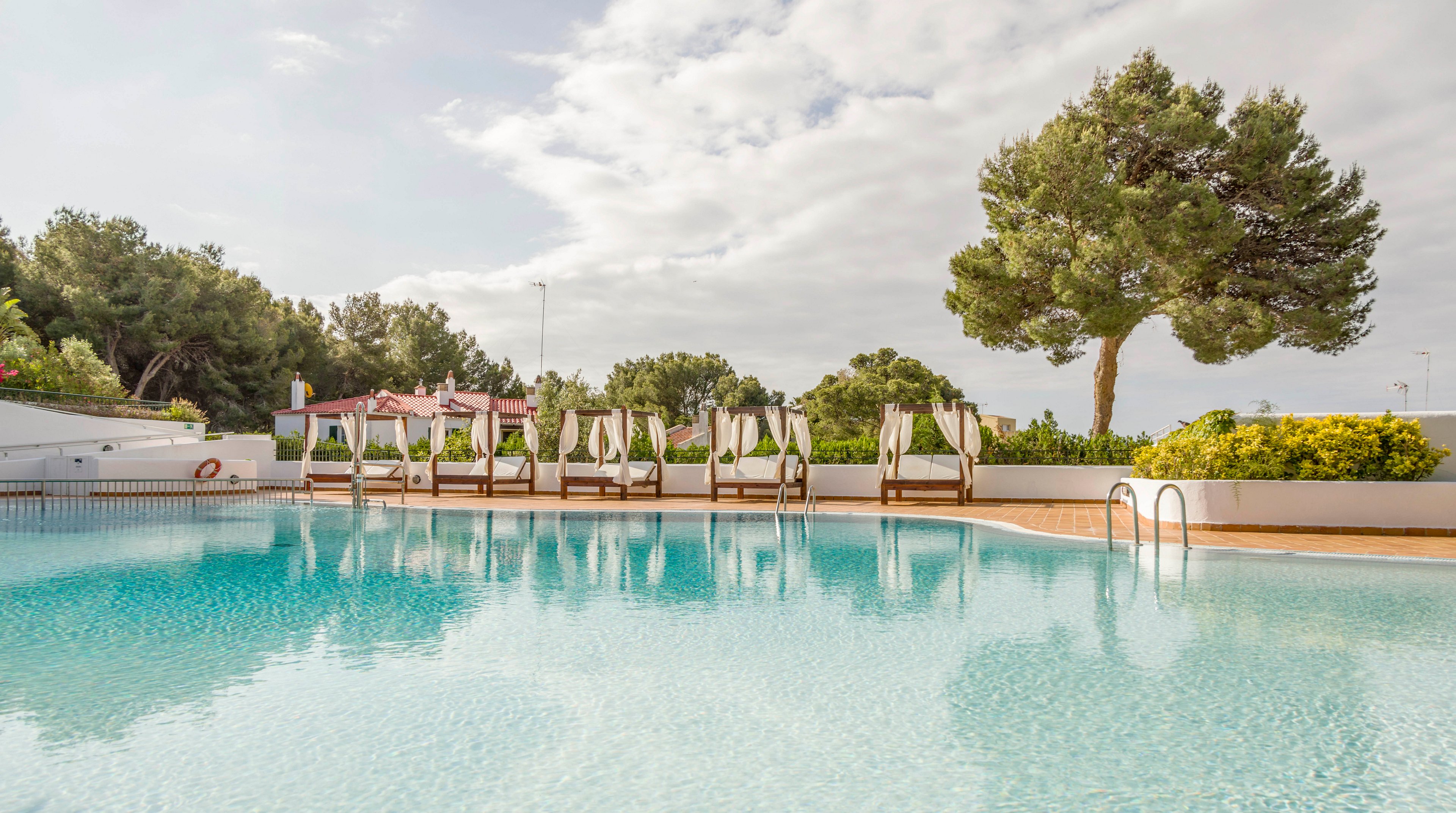 A clear blue swimming pool reflects the bright sky in an outdoor resort setting. Several cushioned loungers with umbrellas line the far side of the pool, inviting relaxation. Tall trees and manicured greenery surround the area, creating a private and tranquil atmosphere. Soft daylight and light clouds contribute to a calm, vacation-like mood.