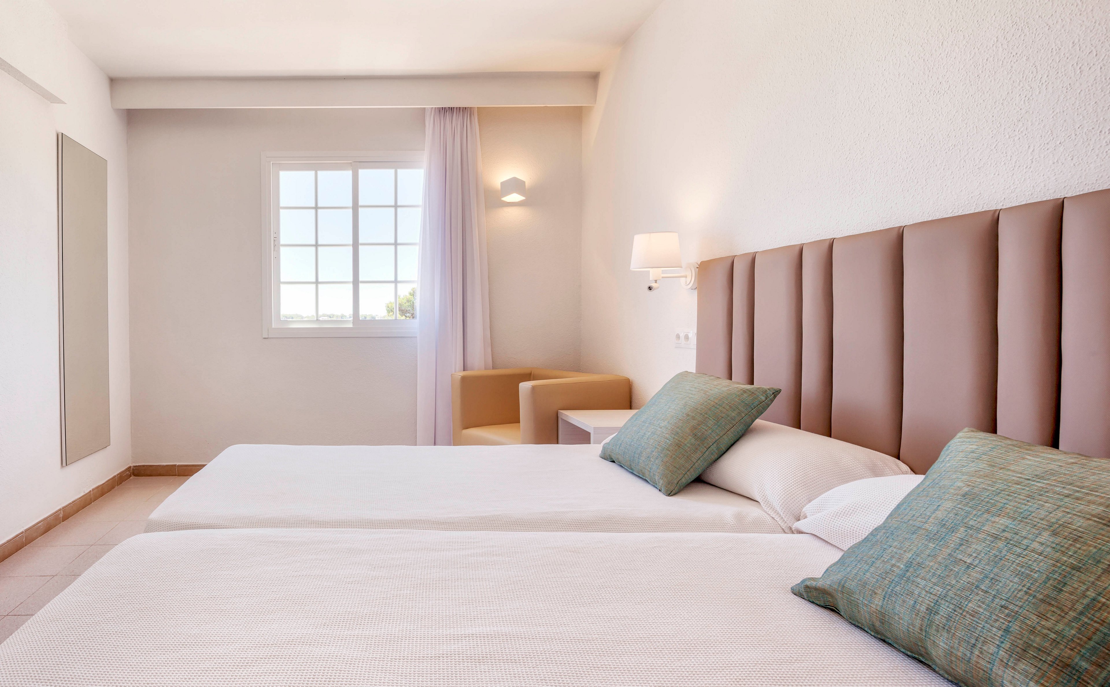 A bright, minimalist bedroom features two neatly made twin beds with padded headboards and pastel cushions. Natural light streams in through a large window with sheer curtains, illuminating the neutral walls and simple decor. A modern wall lamp and a small armchair in the corner add comfort and functionality to the space. The overall atmosphere is calm, clean, and inviting, suitable for a hotel or guest room.