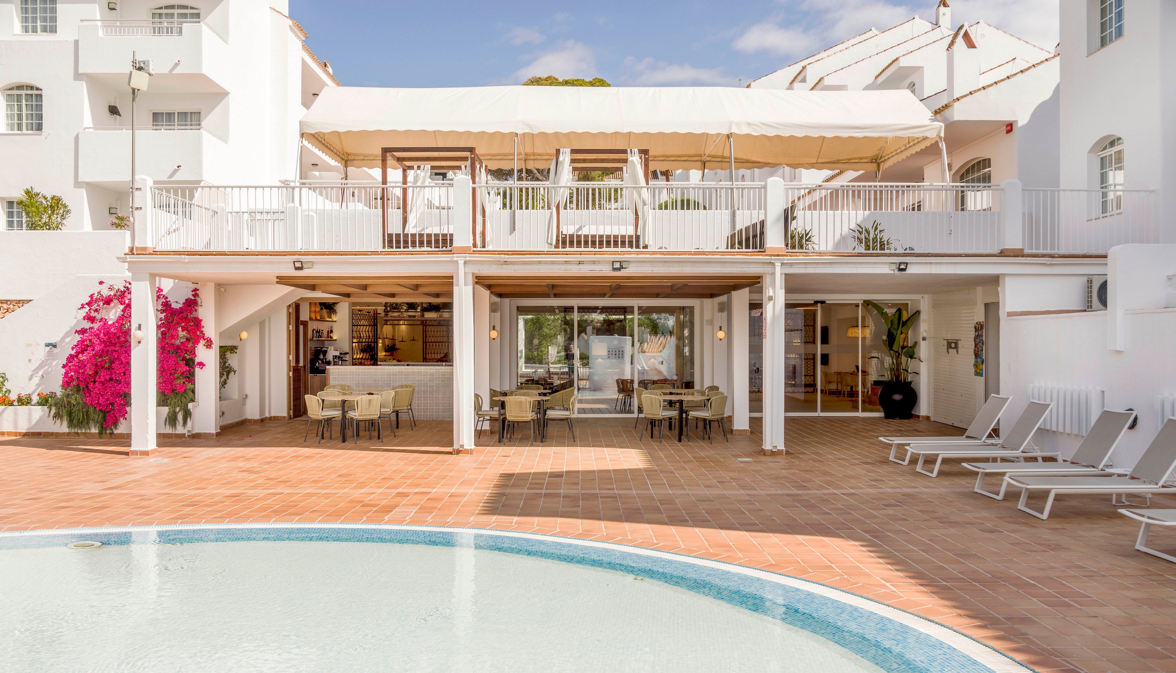 A bright resort building with white walls overlooks a curved swimming pool in the foreground. The scene features an open-air terrace with tables and chairs, and a shaded upper deck area. Several empty sun loungers line the tiled poolside, suggesting a relaxing vacation setting. Pink bougainvillea adds a pop of color against the otherwise neutral architecture under a clear blue sky.