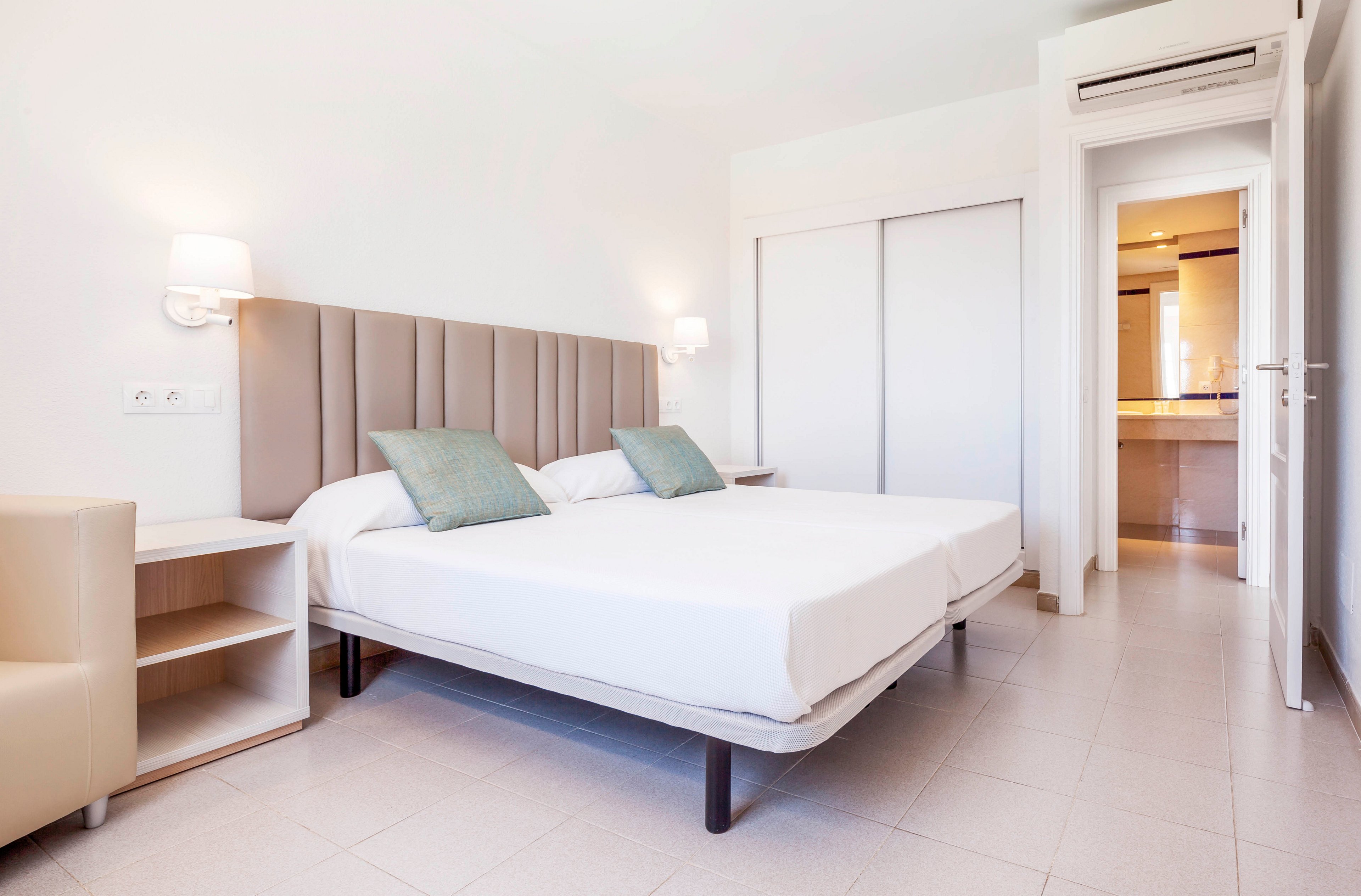 A bright, modern bedroom features two twin beds pushed together with a padded headboard and white linens. Soft wall-mounted lamps and pale tile flooring enhance the clean, minimalist look. Light green accent pillows add a subtle touch of color to the neutral palette. Built-in storage, sliding closet doors, and an open doorway to a lit hallway or kitchenette complete the contemporary hotel-style space.