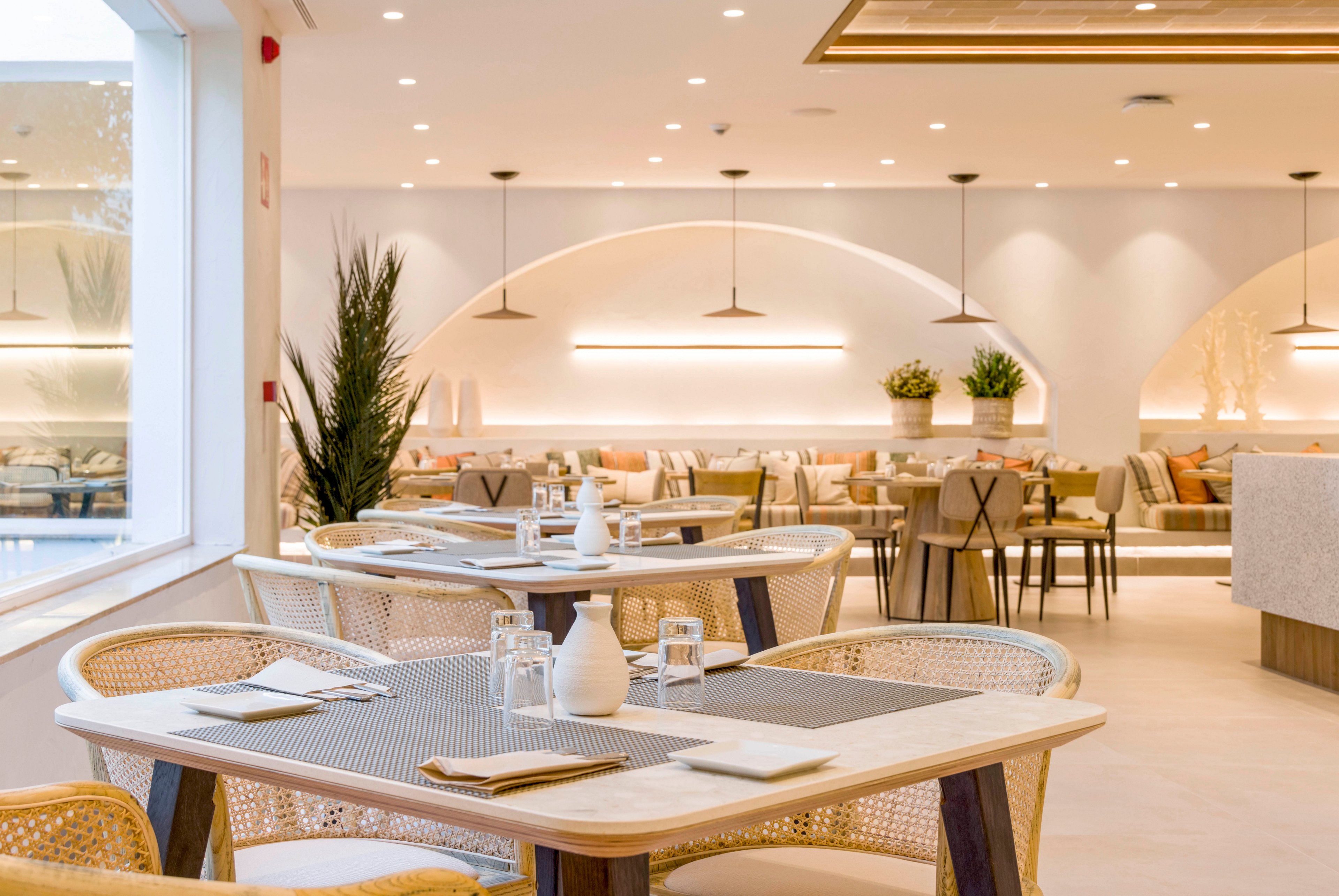A modern restaurant dining area features light wooden tables and woven chairs arranged neatly in the foreground. Soft, warm lighting and minimalist pendant lamps create an inviting atmosphere throughout the spacious room. Neutral tones, indoor plants, and cushioned bench seating along the walls contribute to a clean, contemporary design. Table settings with glasses, plates, and napkins suggest the space is prepared for guests.