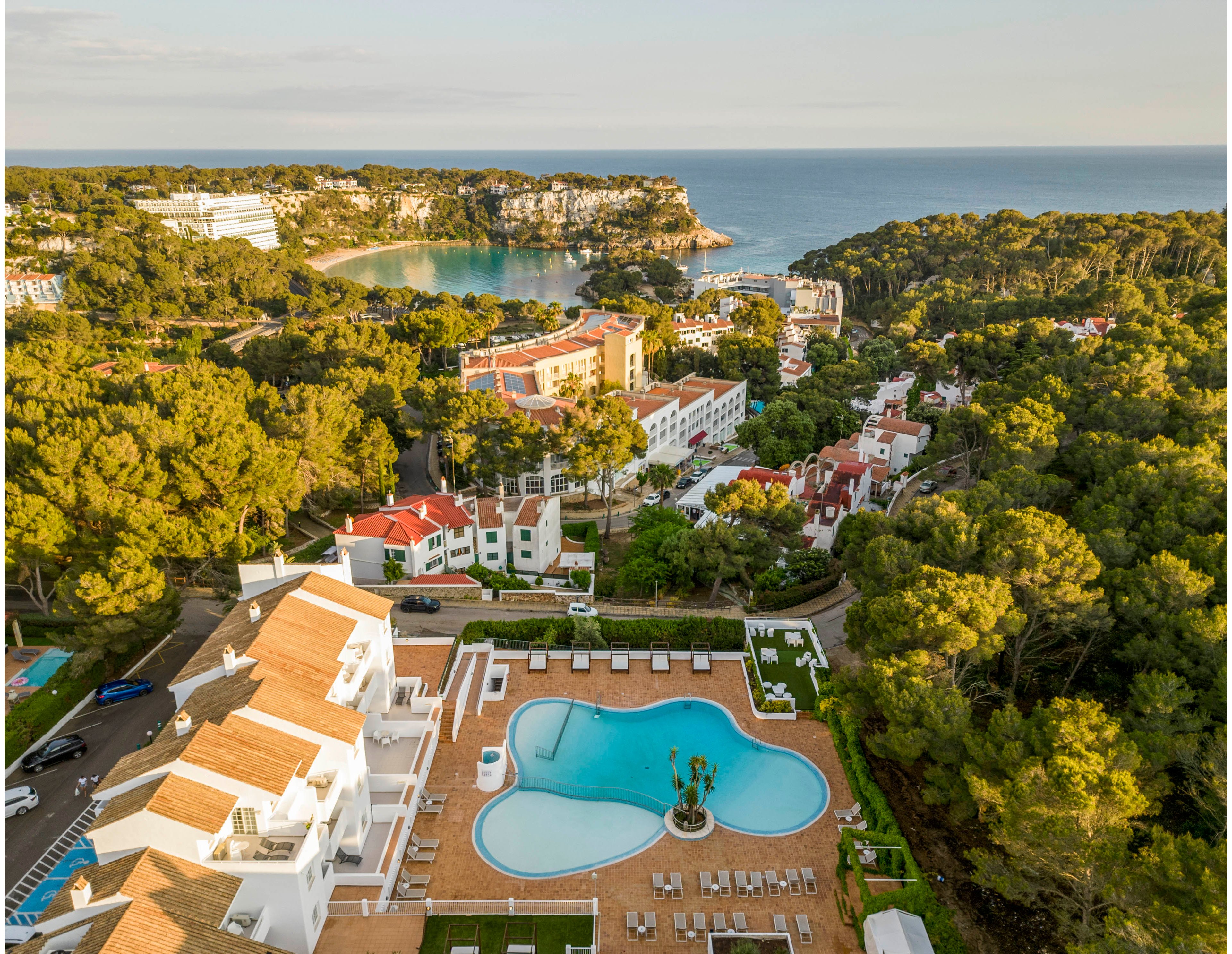 An aerial view captures a coastal resort complex surrounded by dense green trees. In the foreground, a large outdoor swimming pool with a central sculpture is bordered by sun loungers and terracotta-tiled terraces. White buildings with red and beige roofs step down the hillside toward a sheltered bay. In the distance, the calm sea and rugged shoreline complete the scenic holiday destination.