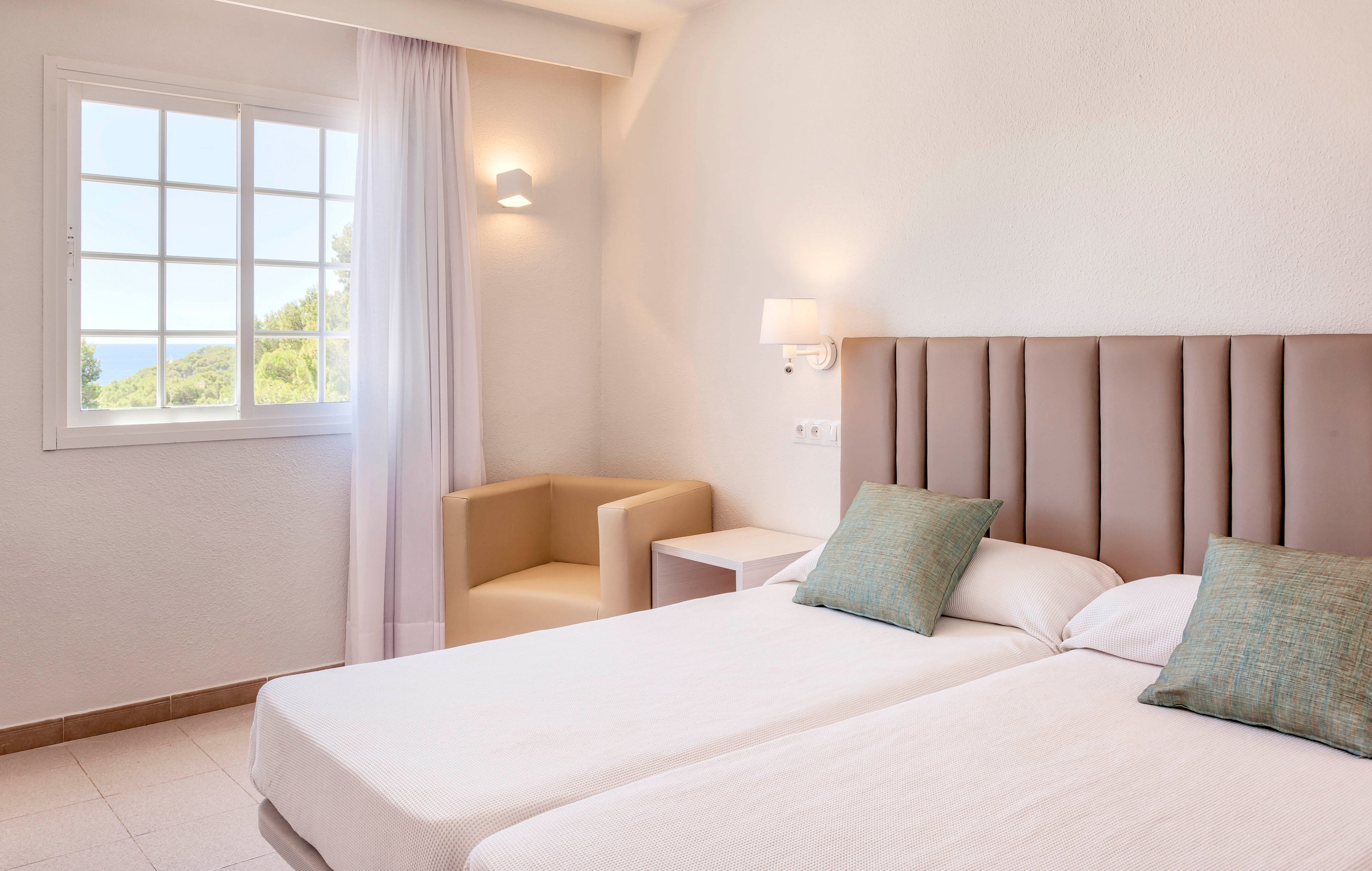 A bright hotel room features two neatly made twin beds with a padded headboard and pastel accent pillows. Natural light streams in through a large window with sheer curtains, revealing a glimpse of greenery and distant water outside. A modern wall lamp, small bedside table, and beige armchair complete the simple, functional layout. The overall design is clean, airy, and contemporary, emphasizing comfort and relaxation.