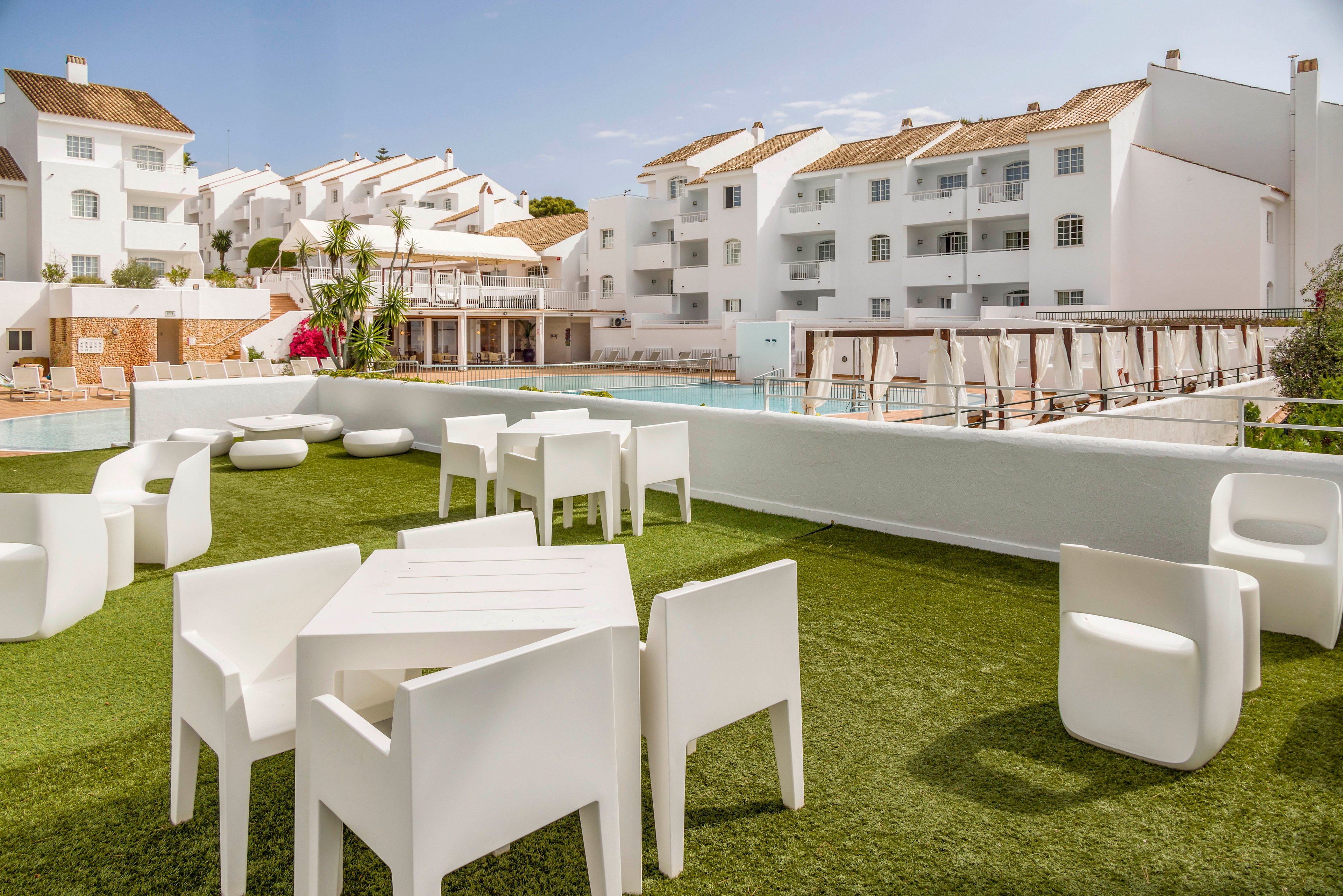 A contemporary outdoor terrace features white minimalist chairs and tables arranged on artificial green grass. Beyond the terrace, a swimming pool stretches alongside a row of white apartment-style hotel buildings. The architecture is clean and modern, with flat facades, balconies, and light-colored tiled roofs. The scene is bright and sunny, creating a relaxed vacation atmosphere in a resort setting.