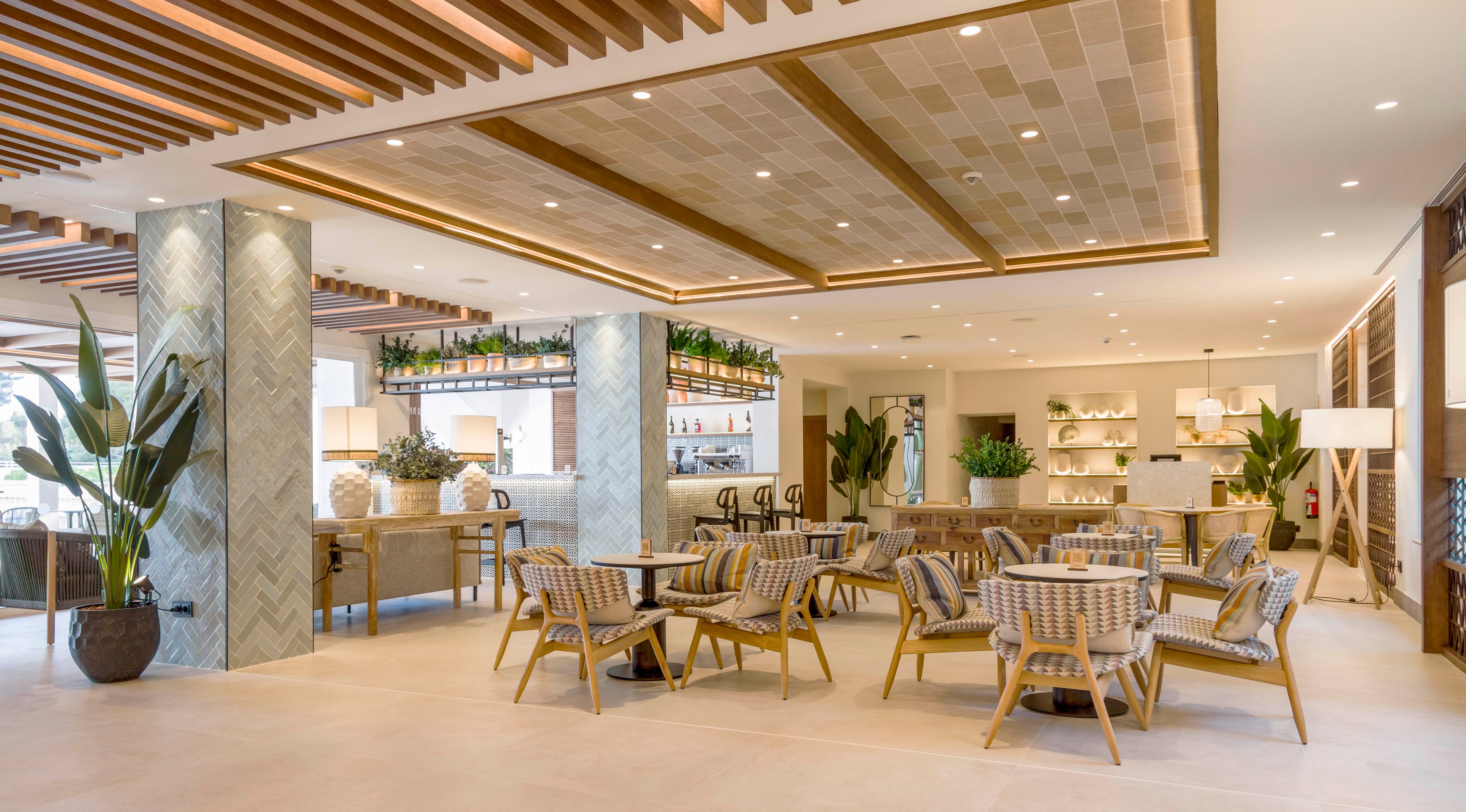 Spacious contemporary lounge area featuring a bar and multiple seating arrangements. Wooden chairs and round tables are arranged across a light tiled floor, with plants adding greenery throughout the space. The ceiling showcases decorative wooden slats and recessed lighting, creating a warm and inviting atmosphere. Shelving behind the bar displays bottles and decor items, emphasizing a stylish hospitality setting.