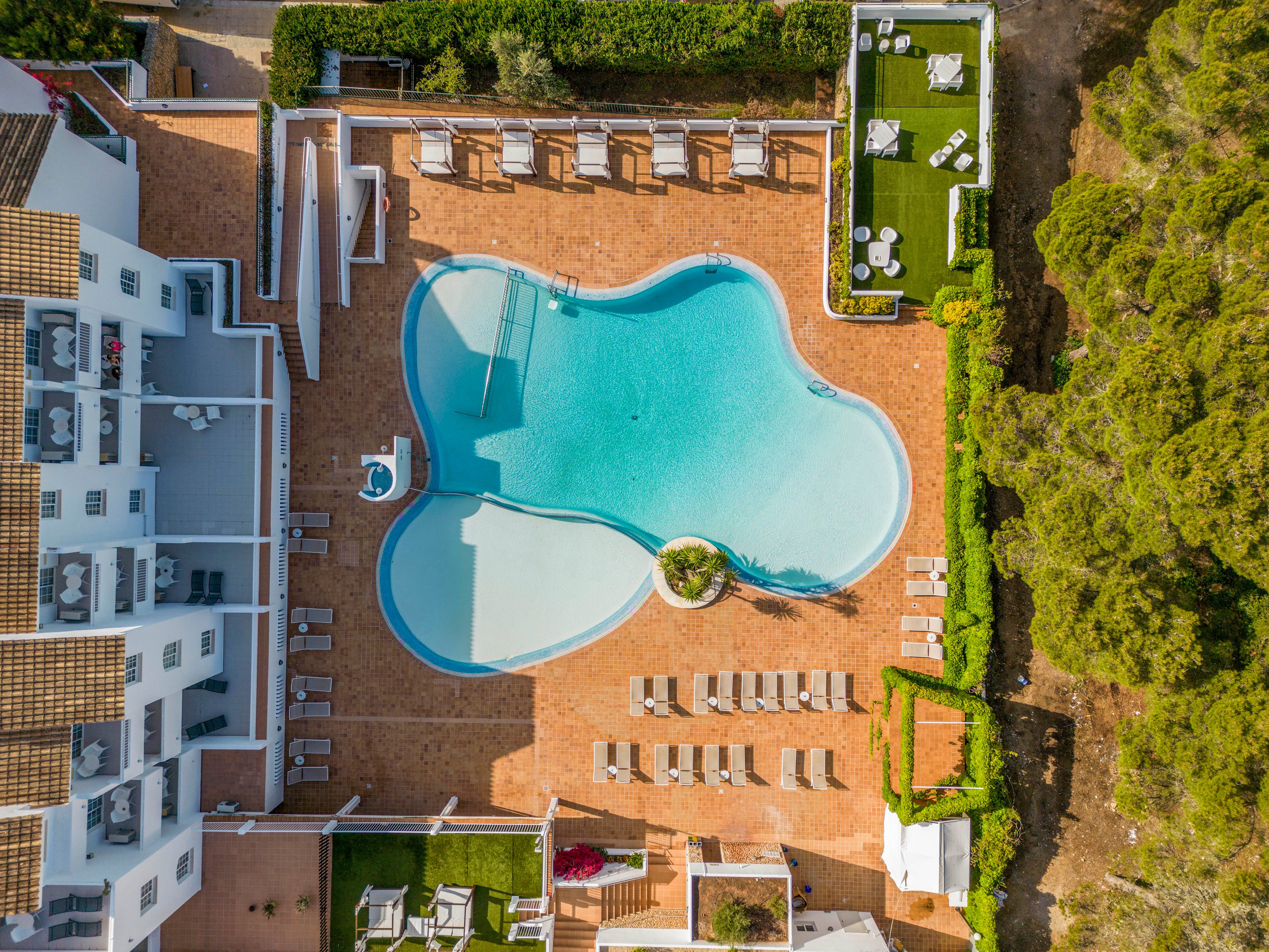 A large outdoor swimming pool is seen from above in the center of a resort complex. Sun loungers and umbrellas line the terracotta-tiled terrace surrounding the pool, with additional seating areas on raised platforms. White buildings with balconies border one side, while green lawns and dense trees frame the other edges. The clear blue water and organized layout create a calm, inviting vacation atmosphere.