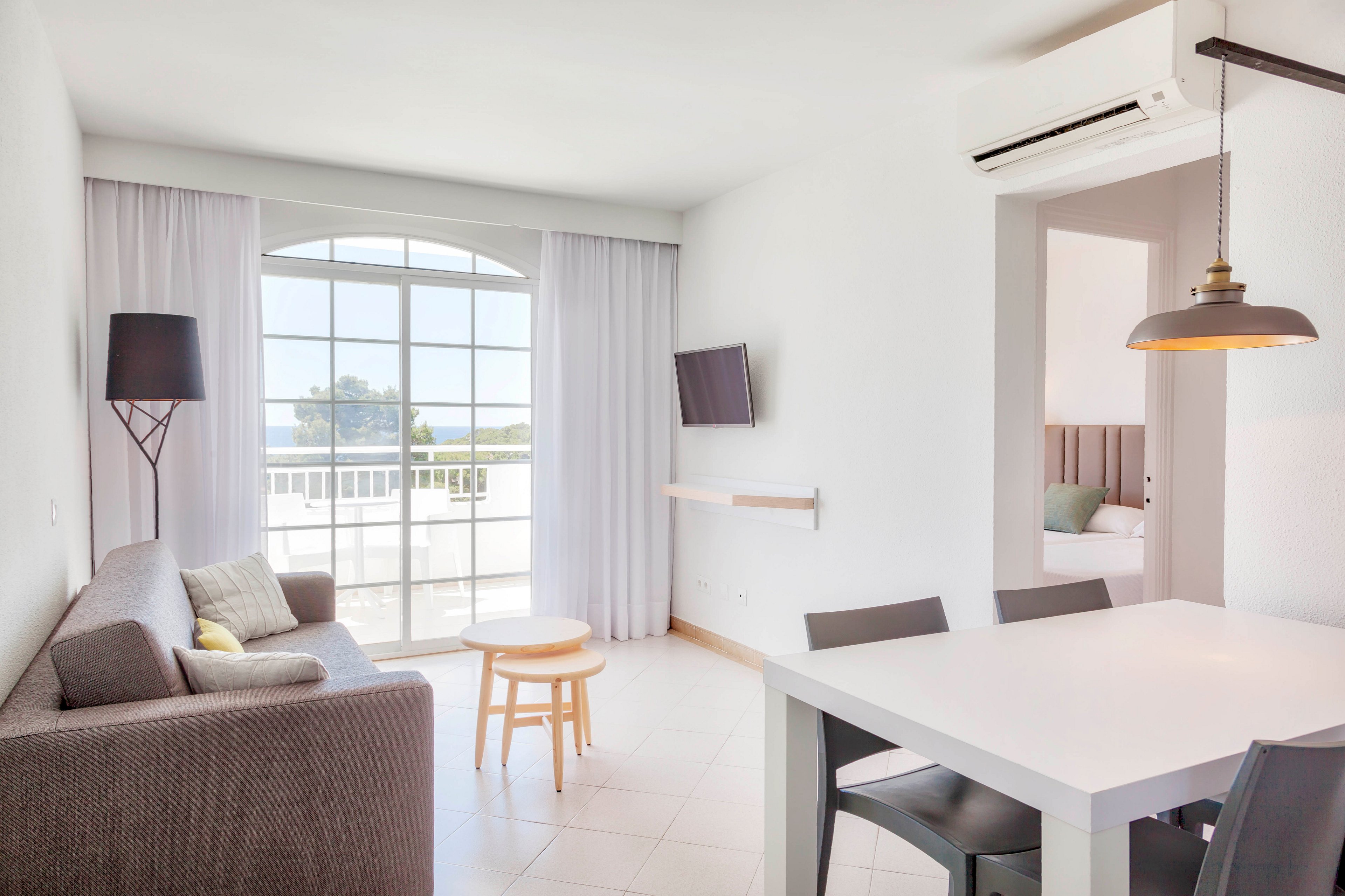 A bright modern hotel suite features a gray sofa, white dining table, and minimalist decor. Large arched windows with sheer curtains open to a balcony and an outdoor view. A wall-mounted television, floor lamp, and small wooden stools add functional touches to the living area. An adjacent bedroom with a neatly made bed is visible through an open doorway, enhancing the spacious, airy feel.