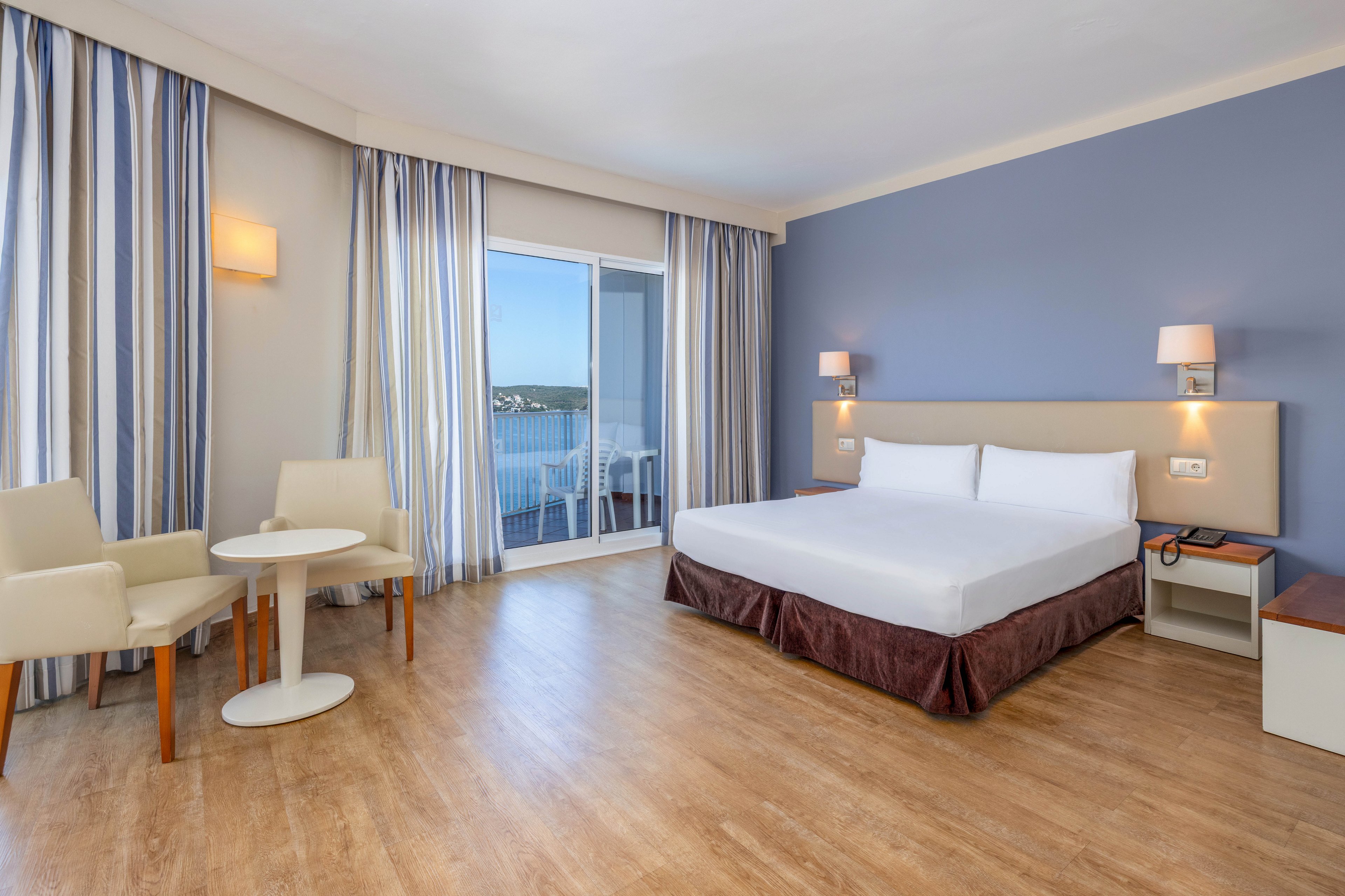 A spacious modern hotel room features a large bed against a blue accent wall. Light wood flooring and neutral furniture create a clean, minimalist look. Floor-to-ceiling curtains frame a sliding glass door that opens to a balcony with a sea view. A small table with two chairs and bedside lamps add comfort and functionality to the space.