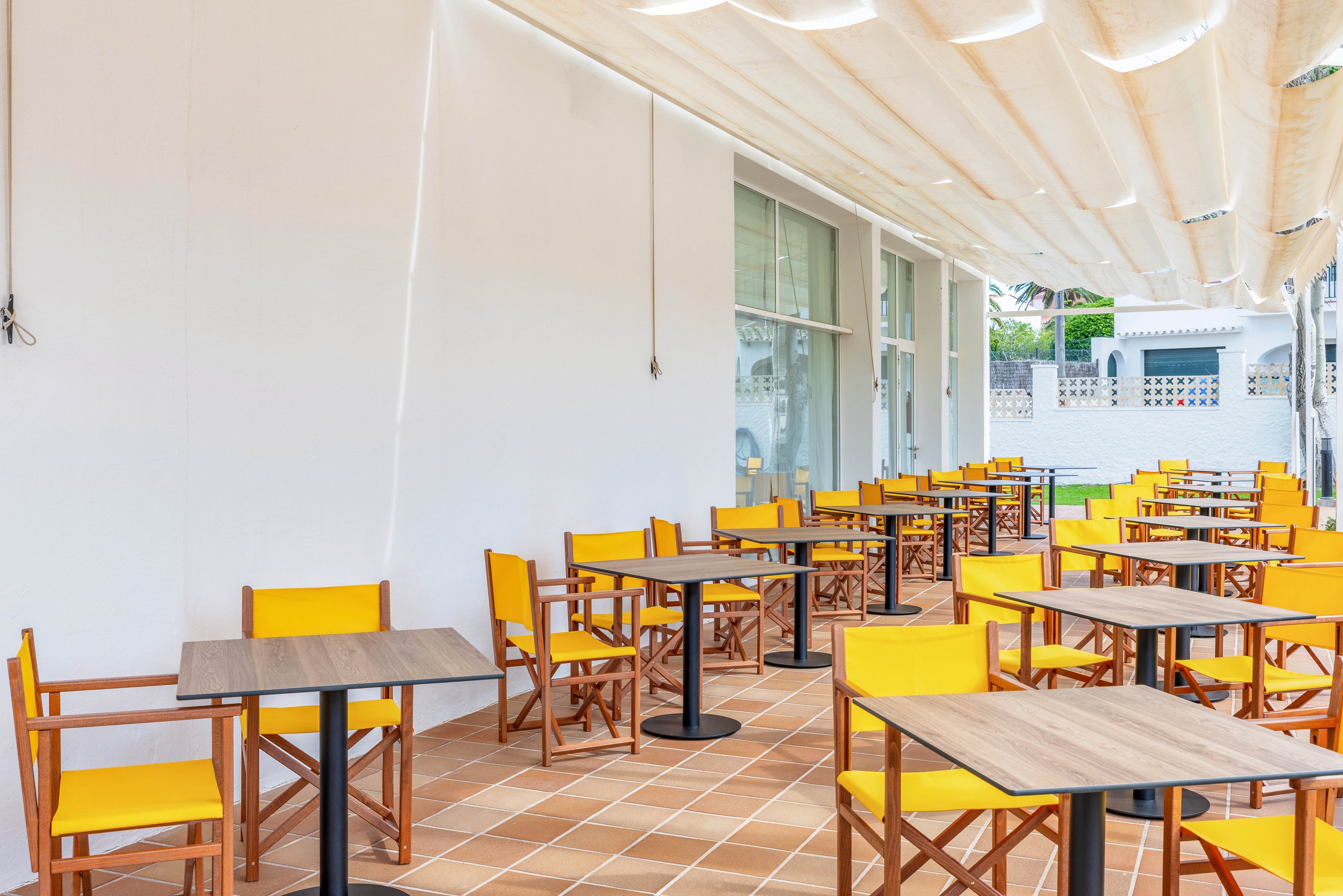 An outdoor café terrace features rows of wooden tables paired with bright yellow chairs. The seating area is arranged on a tiled floor beside a white building with large glass windows. A light-colored fabric canopy provides shade above the tables, creating a comfortable dining atmosphere. The space appears clean, modern, and ready to welcome guests, though no people are present.