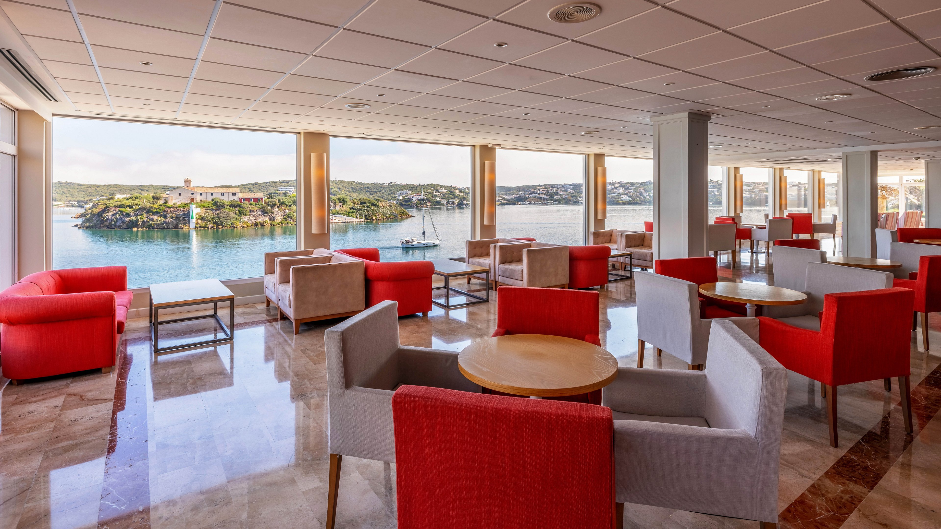 Spacious hotel lounge with contemporary armchairs and small round tables faces large windows with a panoramic view of a coastal bay. Red and beige seating is arranged in conversational groupings on a polished marble floor. Sunlight streams through the floor-to-ceiling windows, illuminating the calm water and distant shoreline outside. The atmosphere feels relaxed, airy, and ideal for quiet conversation or enjoying the scenery.