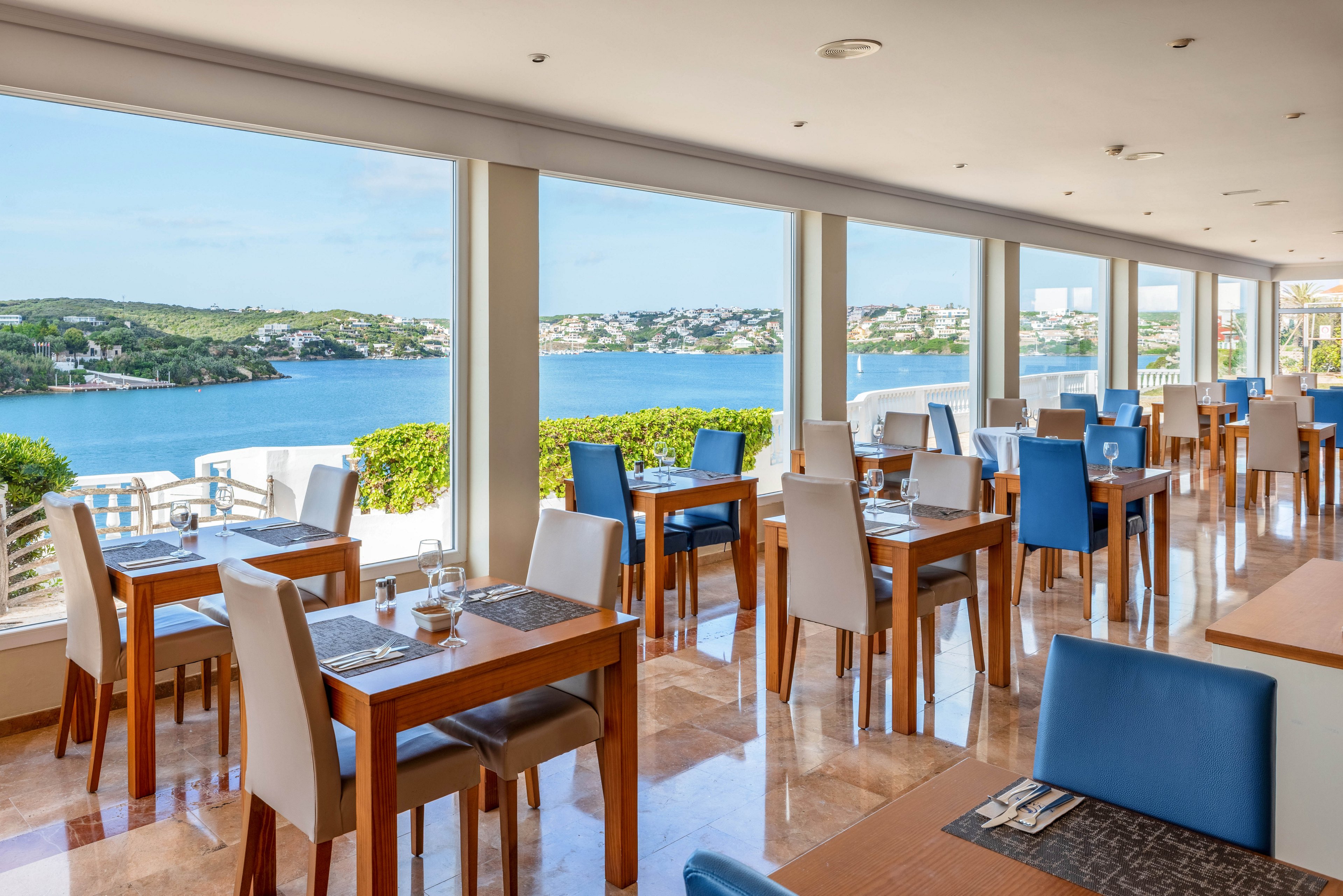 Contemporary dining tables and chairs are arranged in a bright waterfront restaurant overlooking a calm bay. Large floor-to-ceiling windows frame expansive views of blue water, green hills, and distant buildings. Sunlight reflects off the polished floor, enhancing the airy and spacious atmosphere. Table settings with placemats, glasses, and cutlery suggest a relaxed yet refined dining experience by the sea.