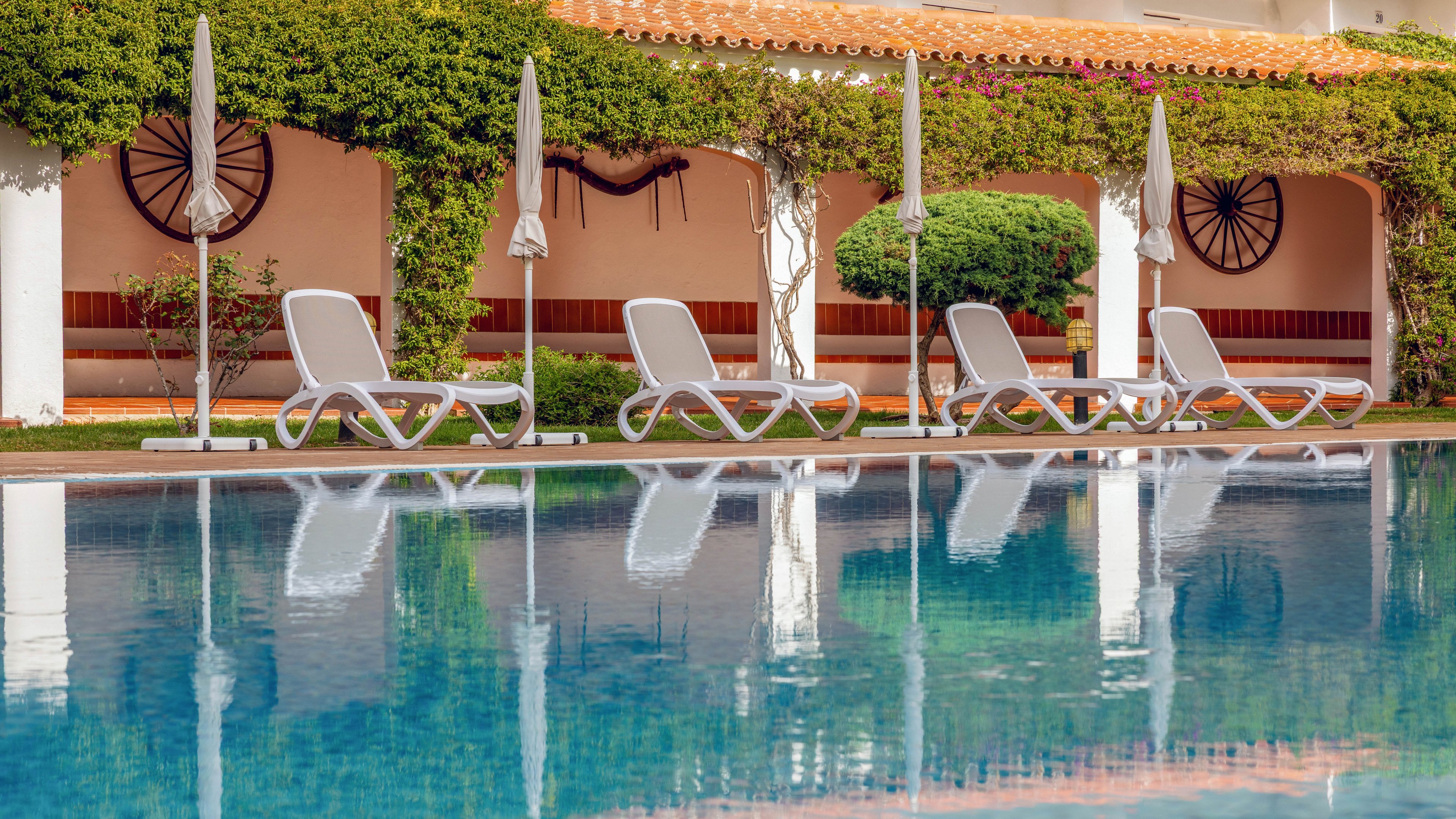A tranquil outdoor swimming pool reflects a row of white sun loungers and closed umbrellas. The loungers are arranged on a poolside terrace in front of a building covered with lush green vines. Decorative wagon wheels hang on the wall, adding a rustic touch to the resort setting. The calm water and soft colors create a peaceful, relaxing vacation atmosphere.
