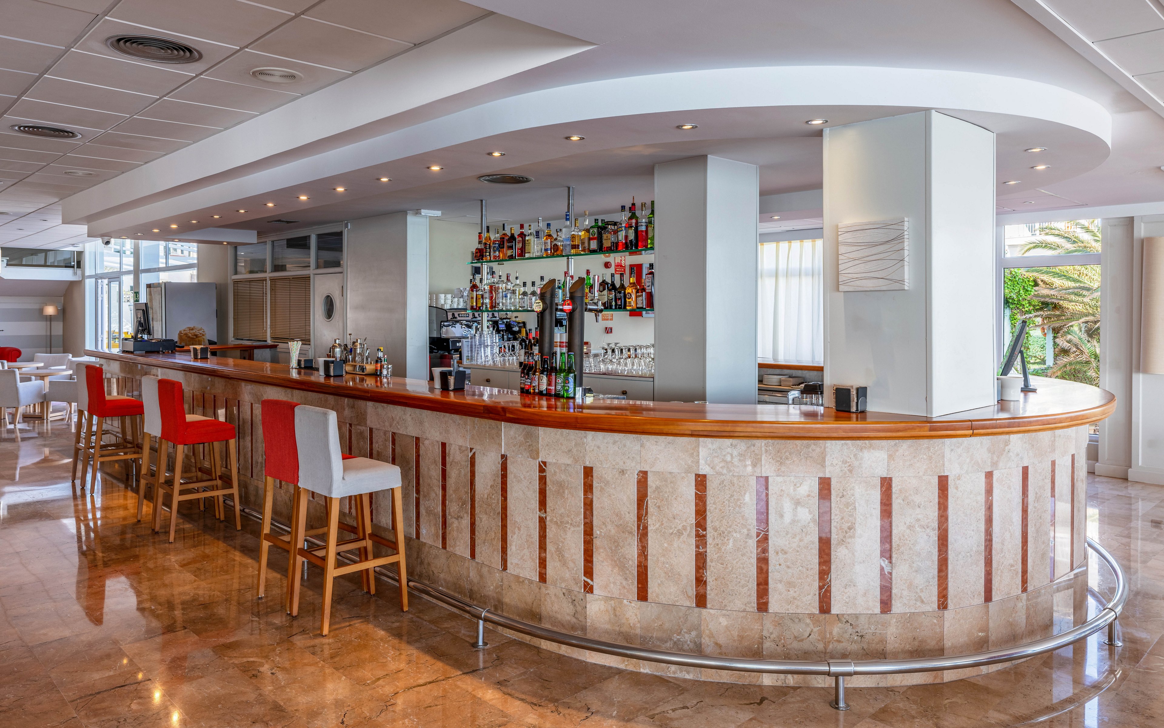 A curved marble-topped bar counter stands in the center of a bright, modern hotel lounge. Colorful bottles and glassware are neatly arranged on shelves behind the bar, with several drink dispensers and equipment on the counter. Tall bar stools with red and white upholstery line the front of the bar, reflecting on the polished floor. Large windows and light-colored walls create an airy, inviting atmosphere, while the space remains empty of people.