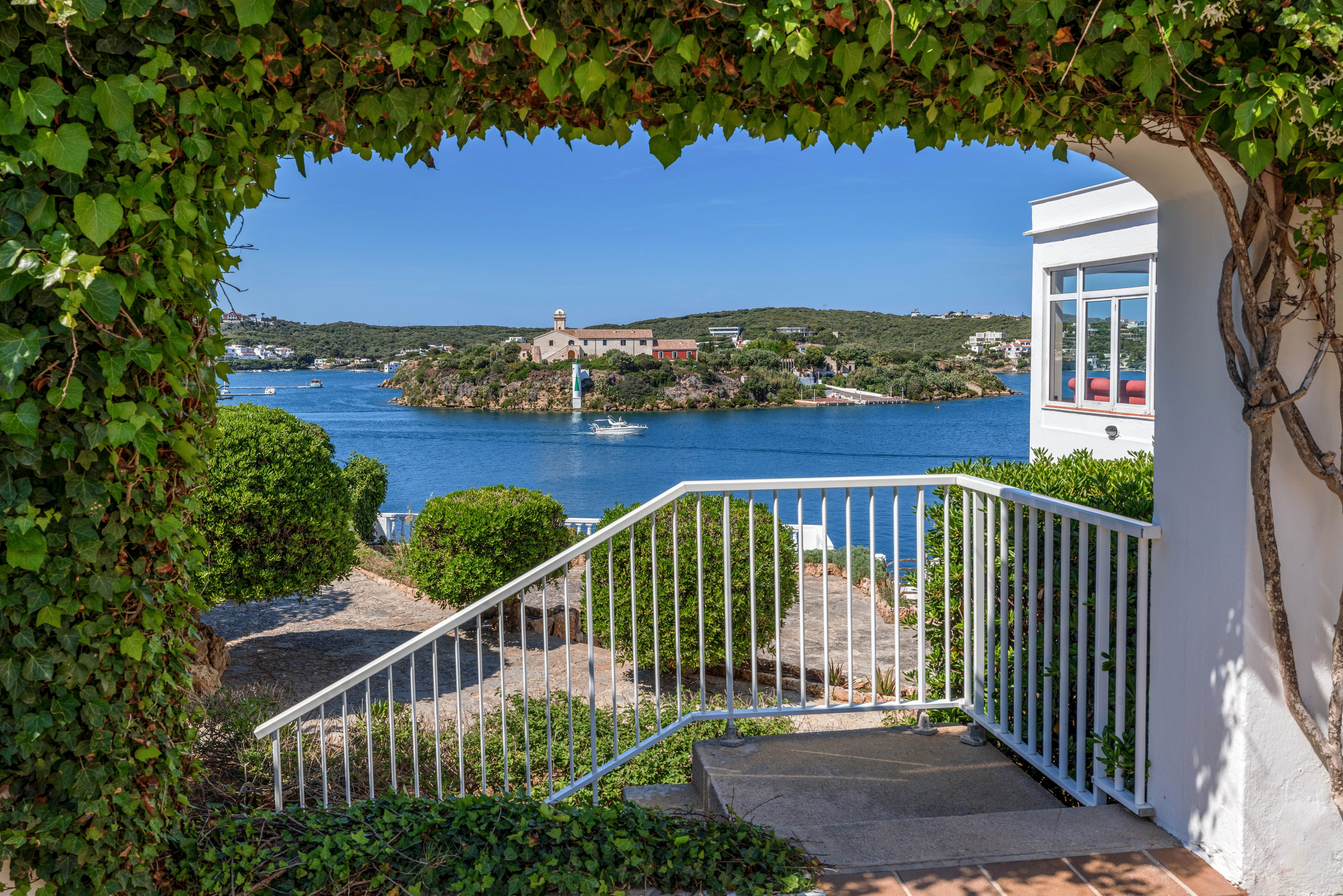 A white terrace framed by lush green ivy opens onto a view of a calm blue bay. Across the water, a small coastal village with a church and scattered houses sits on a low hill. Boats are visible on the water, enhancing the leisurely seaside atmosphere. Bright sunlight and clear skies create a warm, inviting Mediterranean feel.