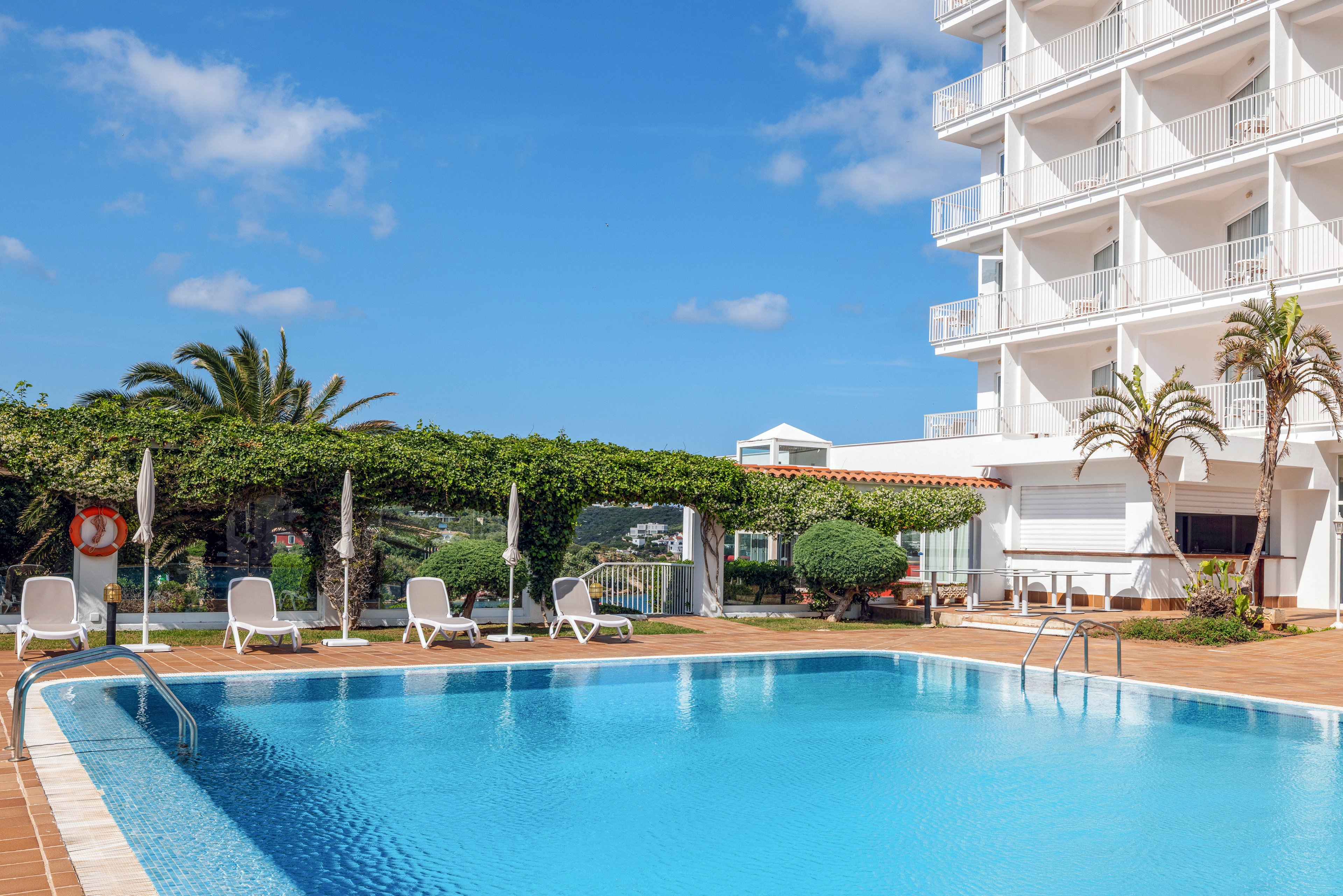 An outdoor swimming pool sits beside a modern white hotel building under a clear blue sky. White sun loungers and umbrellas line the pool deck, creating a relaxing resort atmosphere. Palm trees and trimmed greenery surround the area, adding a tropical touch. The calm water and bright daylight suggest a warm, inviting vacation setting.