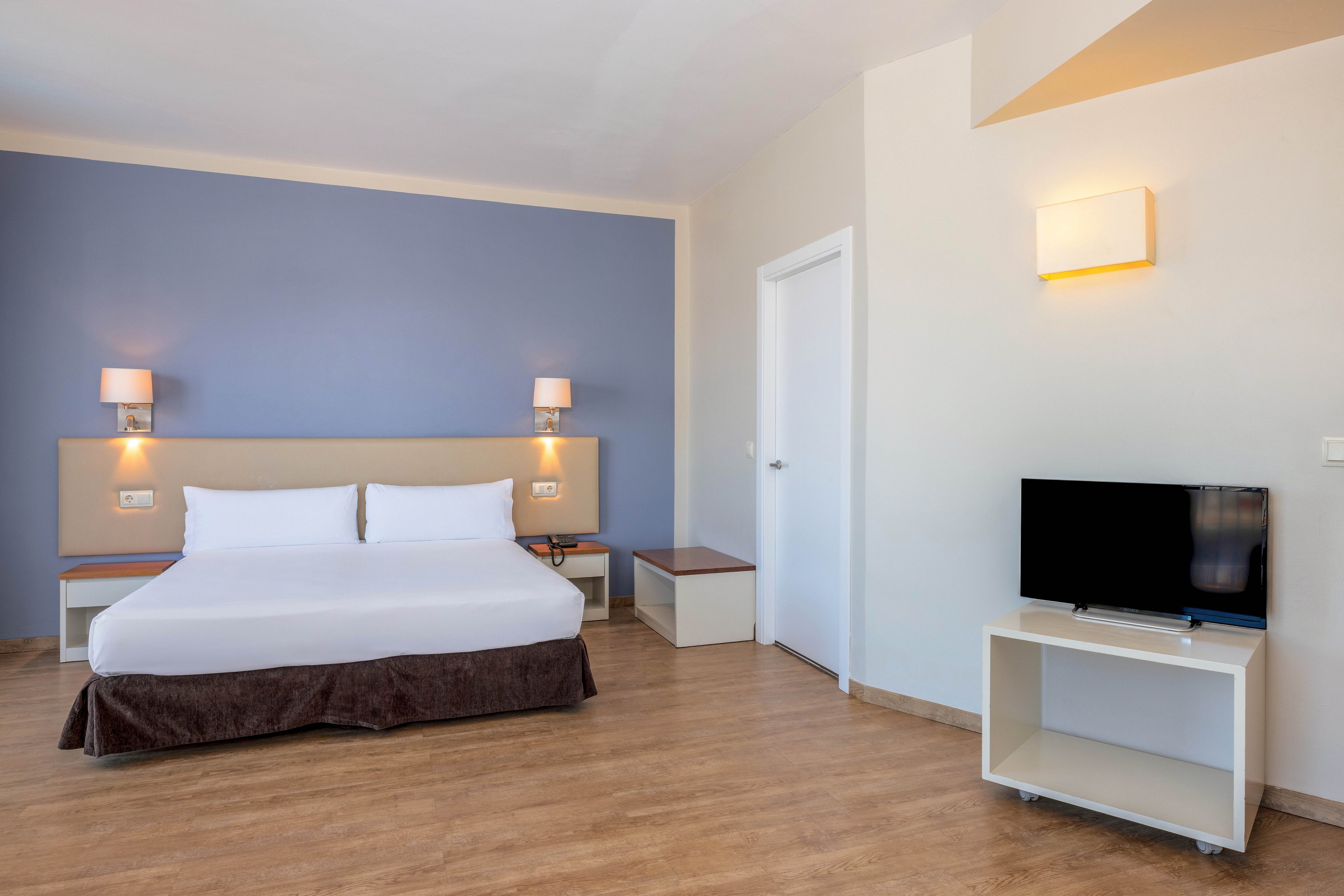 A modern hotel room features a large neatly made bed against a blue accent wall. Two wall-mounted lamps flank the headboard, providing warm ambient lighting. On the right side of the room, a flat-screen television sits on a white stand with open shelving. The space has light-colored walls, wood flooring, and a simple door, creating a clean and minimalist atmosphere.